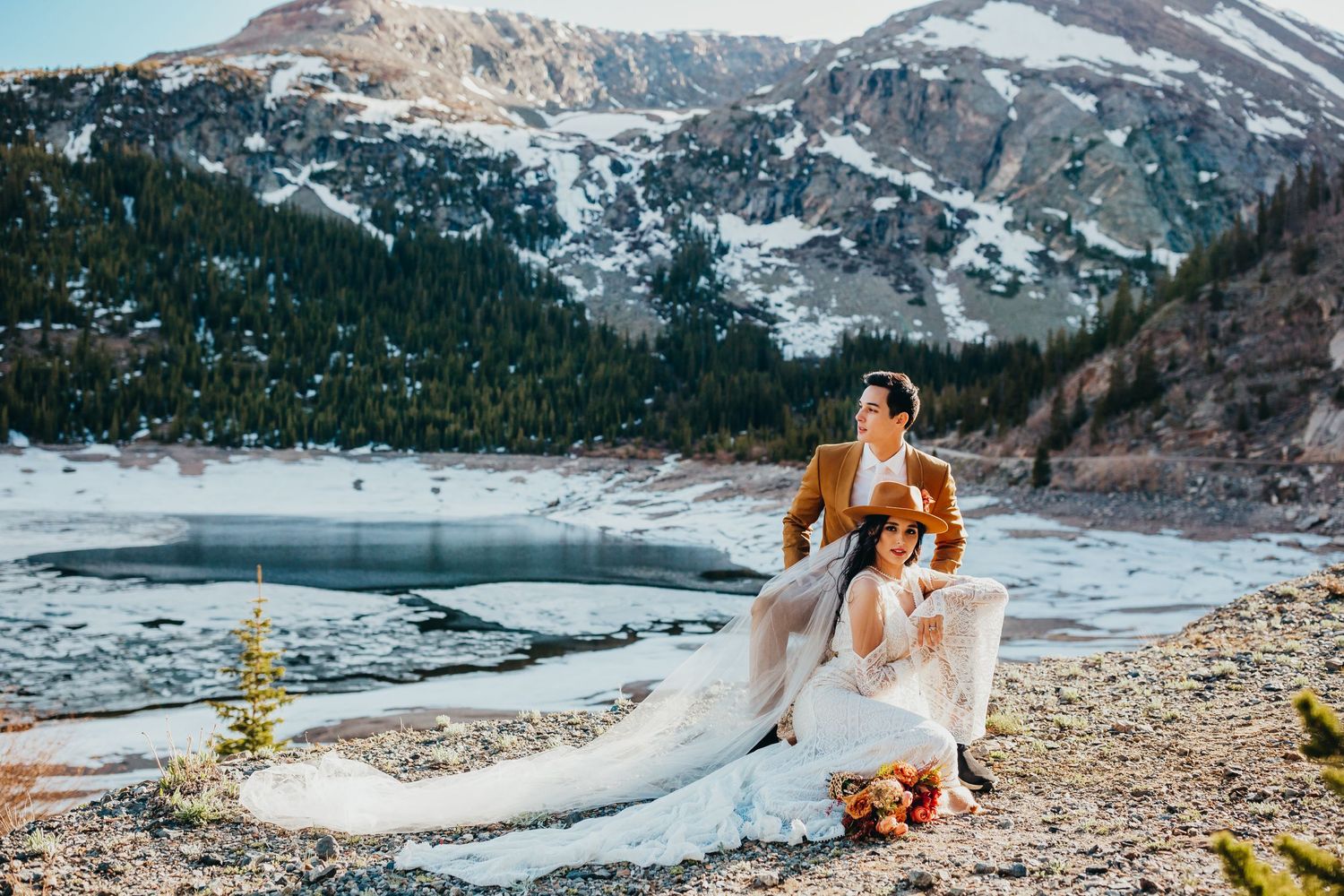 A flowing white wedding dress billows in the wind against a snowy mountain landscape beside an icy lake.