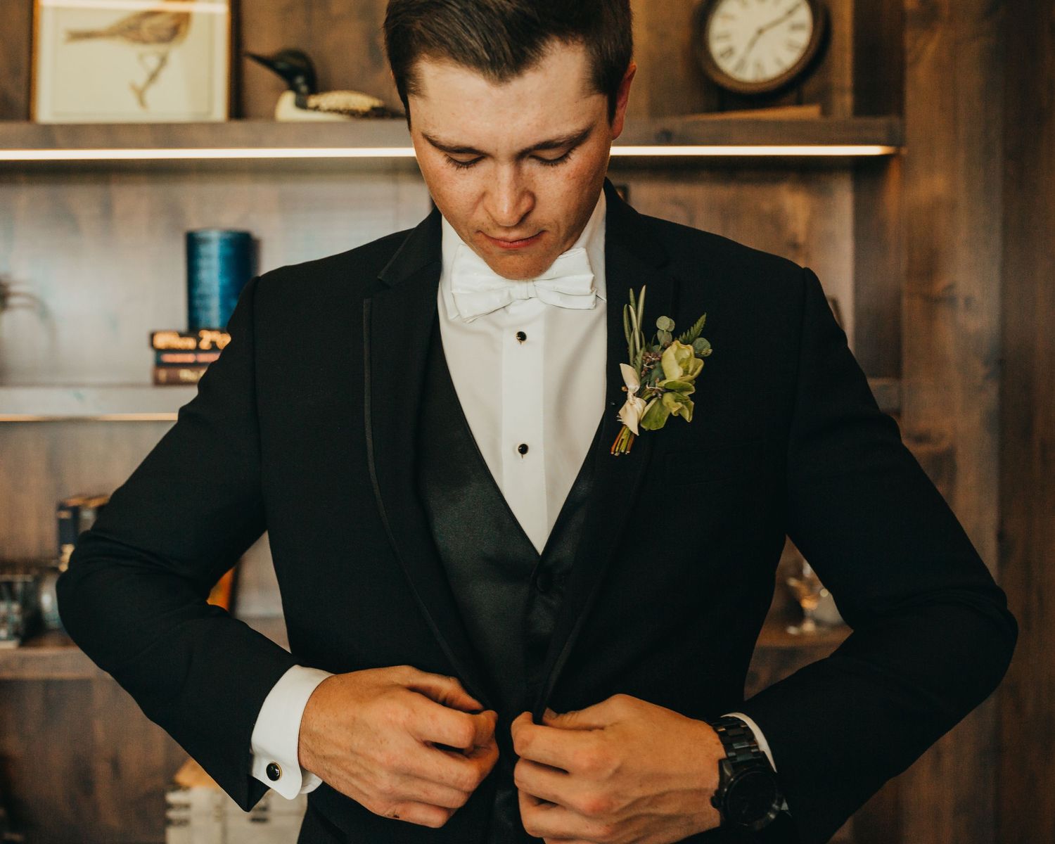 A groom adjusts his black suit jacket while wearing a white bowtie and boutonniere.