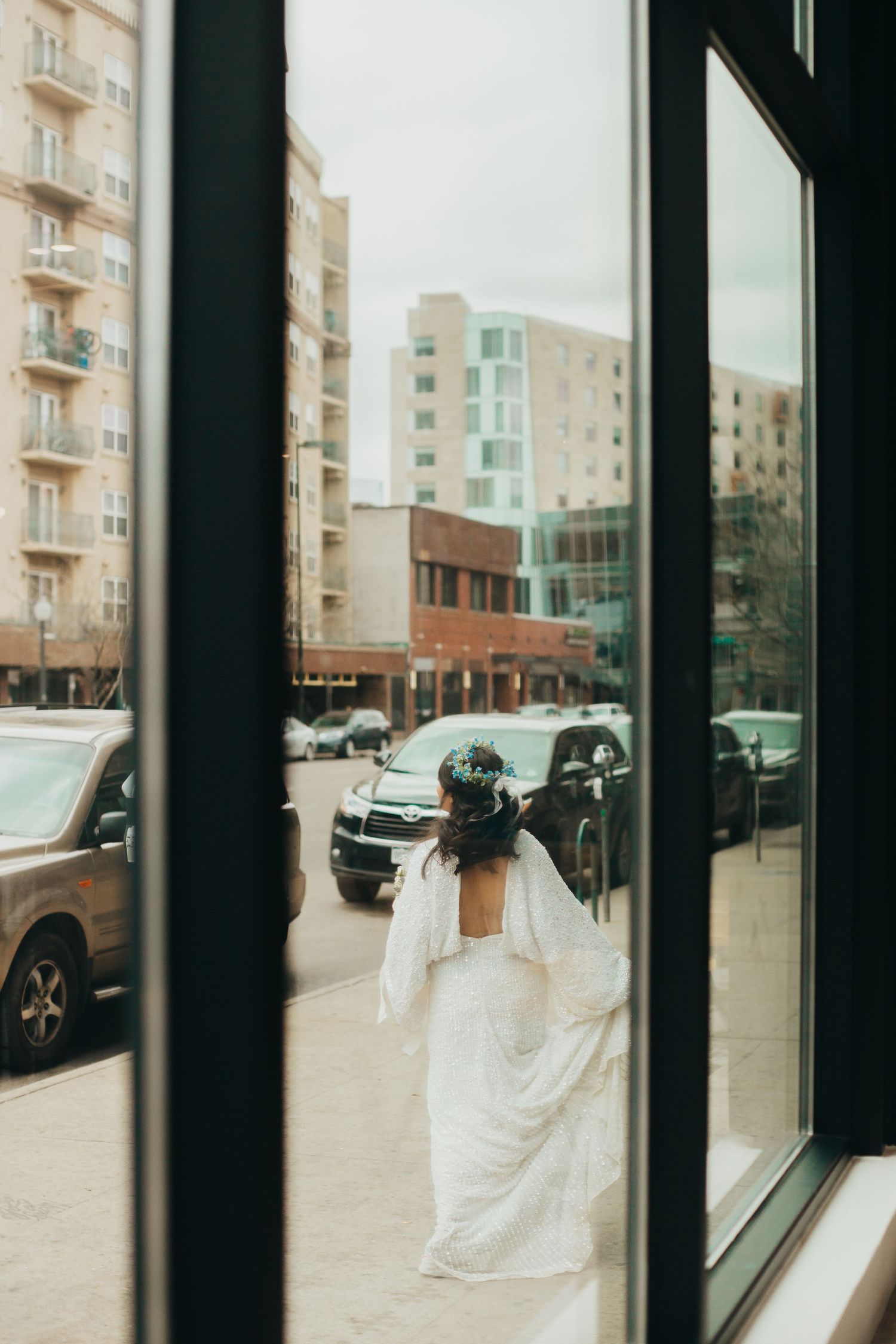 A bride in white dress seen through a window looking at city buildings.