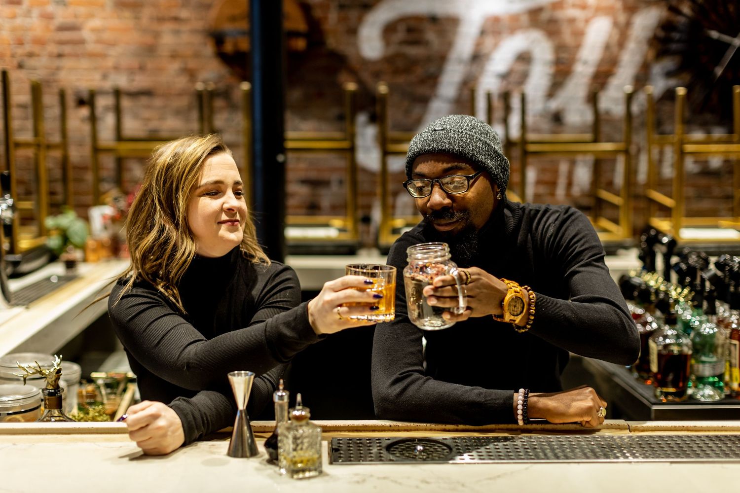 Two friends enjoying drinks together at a rustic bar counter with brick walls in the background.