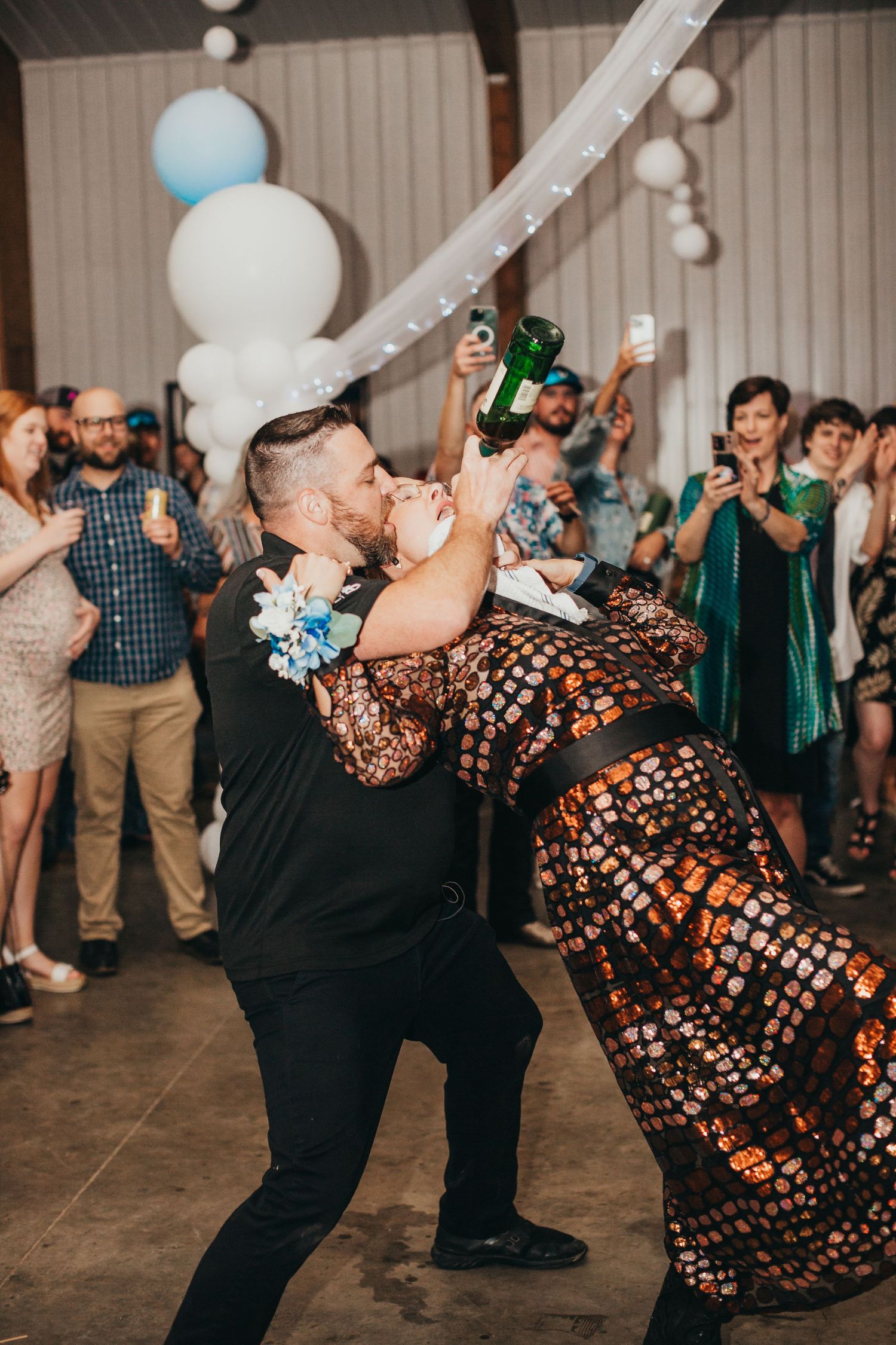 Couple performs fun dip dance move at wedding reception with crowd cheering in background with festive decorations.