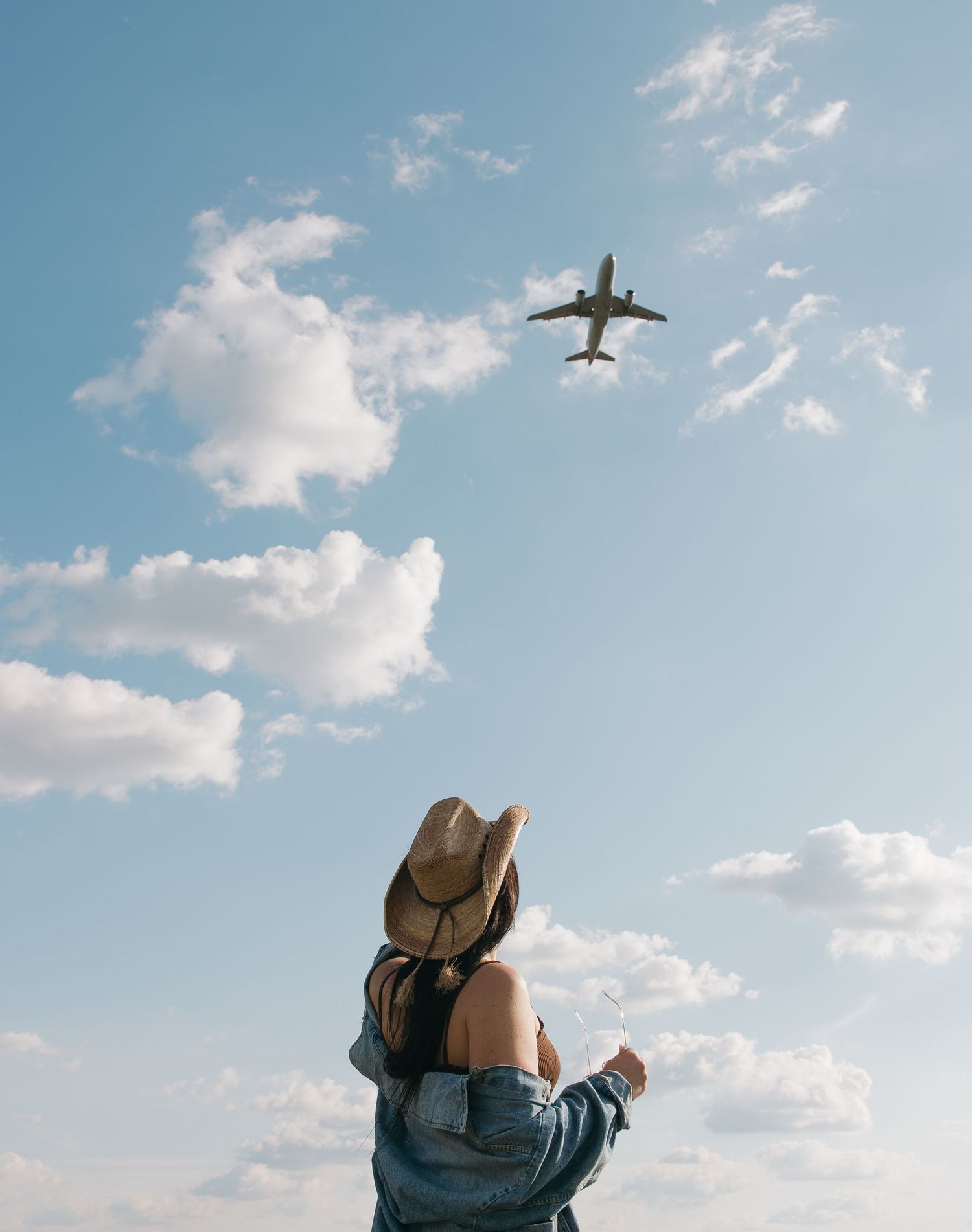 A person in a denim jacket and hat watches an airplane fly overhead on a sunny day. Sioux Falls, South Dakota.
