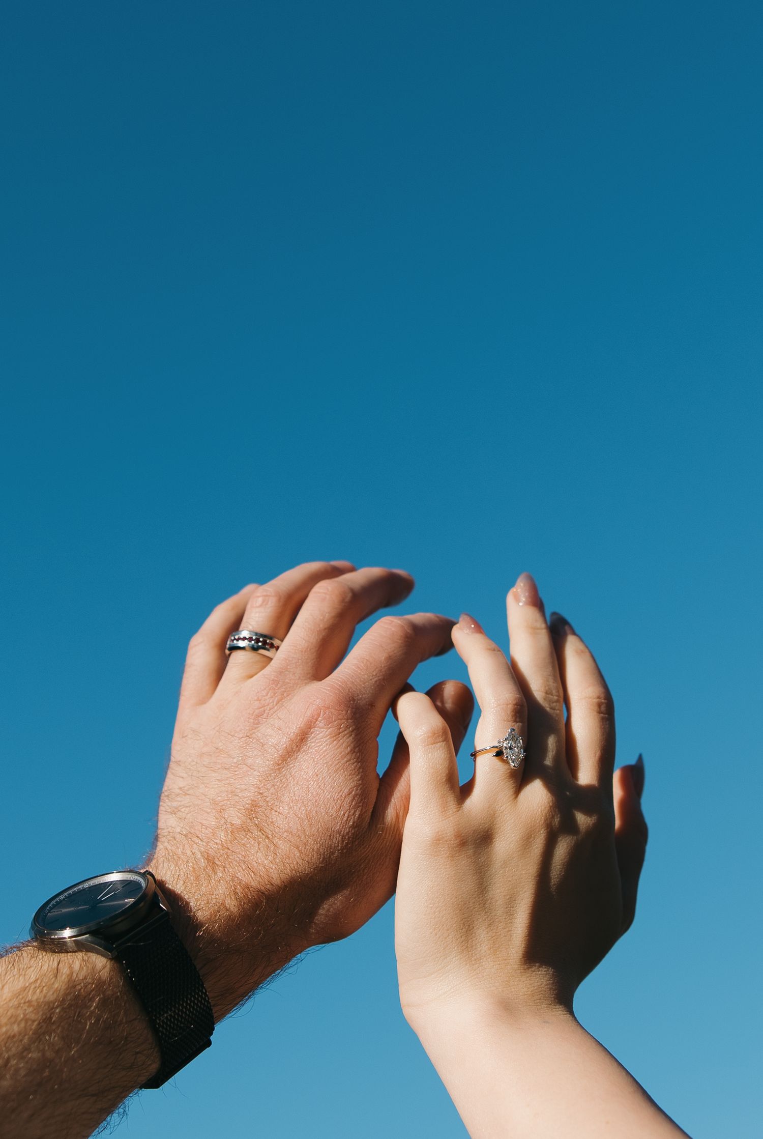 Two hands raised against blue sky, wearing matching rings, with a watch on the man's wrist.