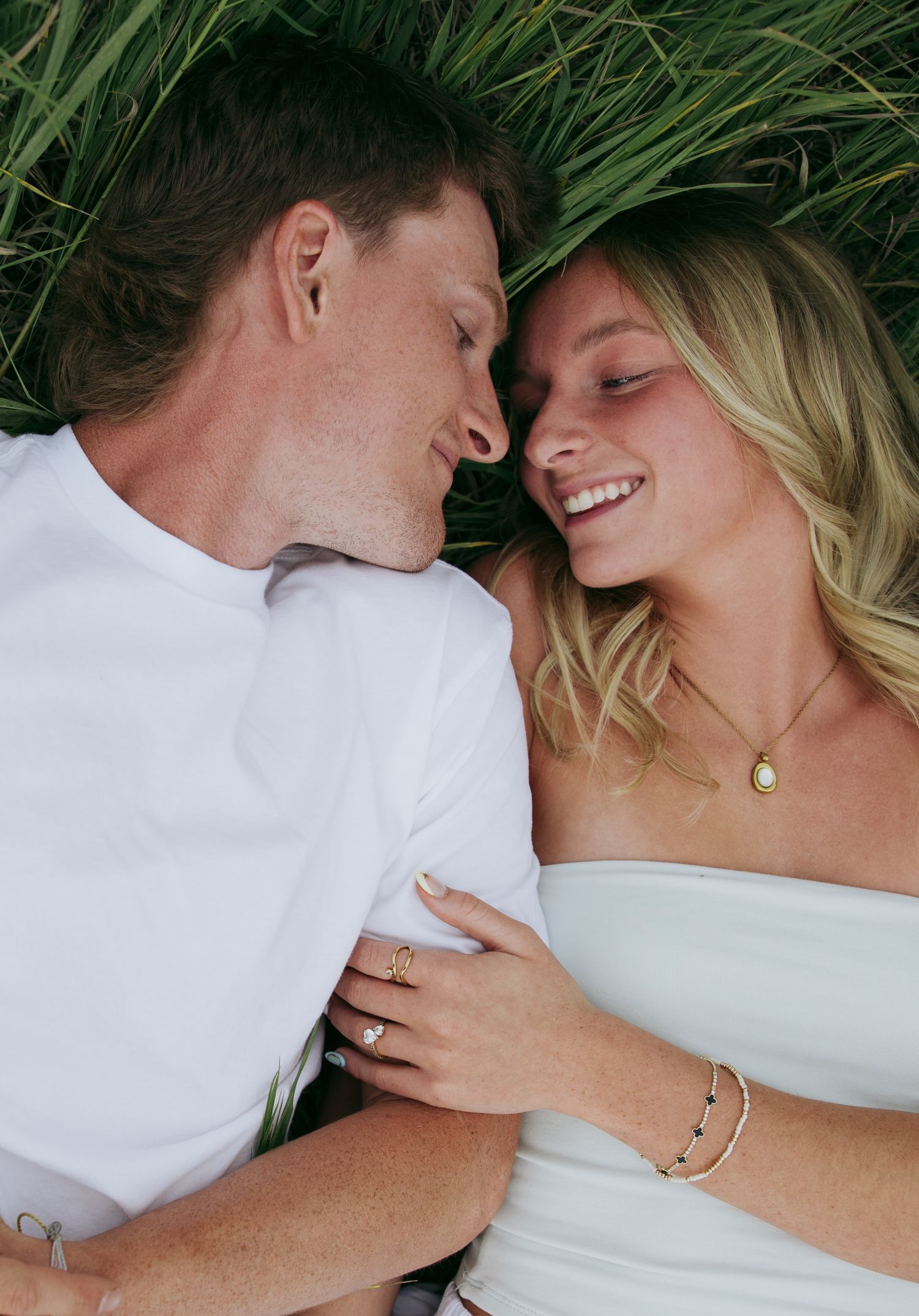 Couple lying in green grass, smiling and gazing at each other, wearing white outfits and gold jewelry.