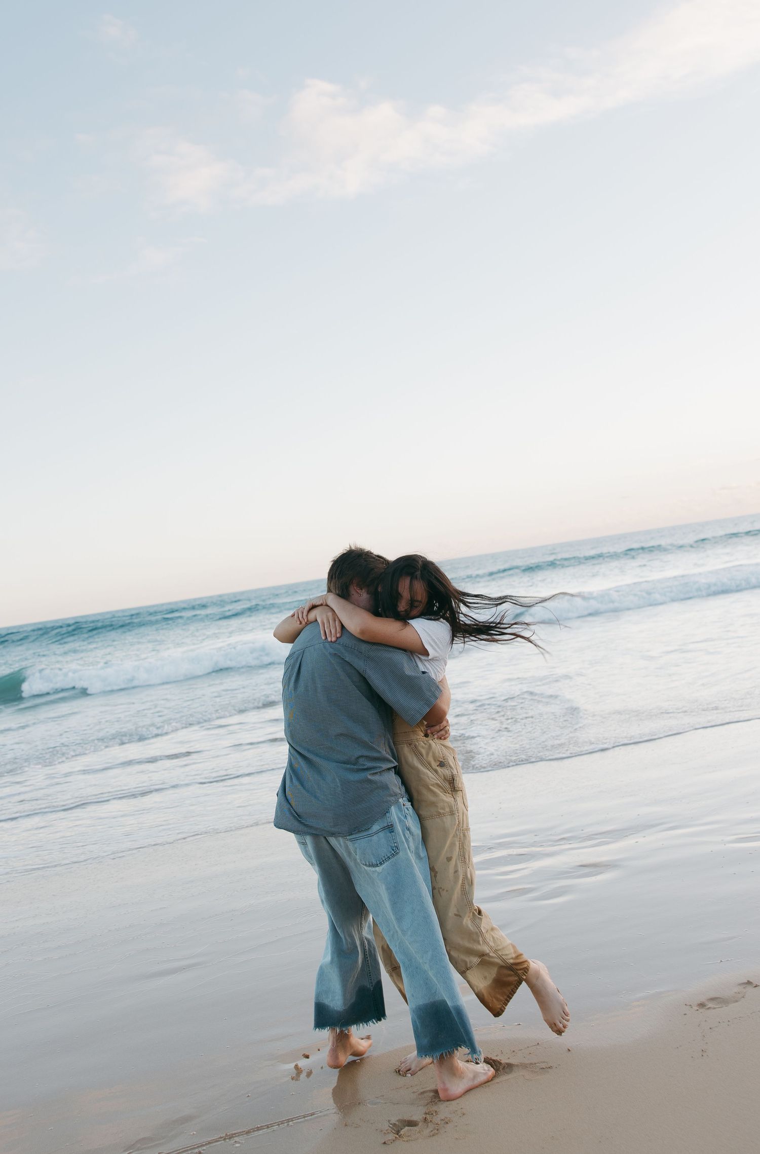 A couple embraces while dancing barefoot on a beach with ocean waves in the background. Sacramento, California. Newport Beach, California.