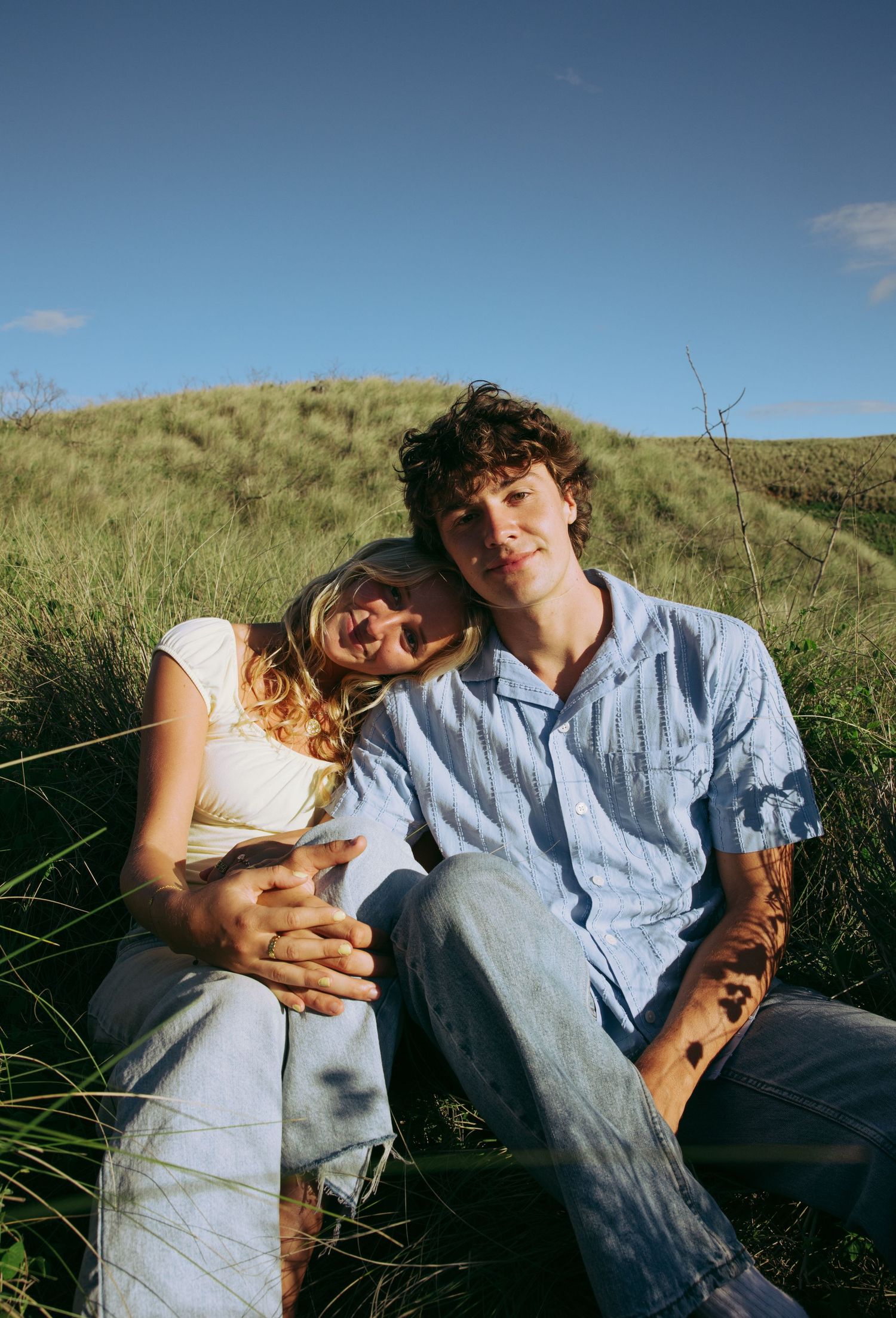 A young couple sits together in a green grassy field under a blue sky, smiling warmly at the camera.