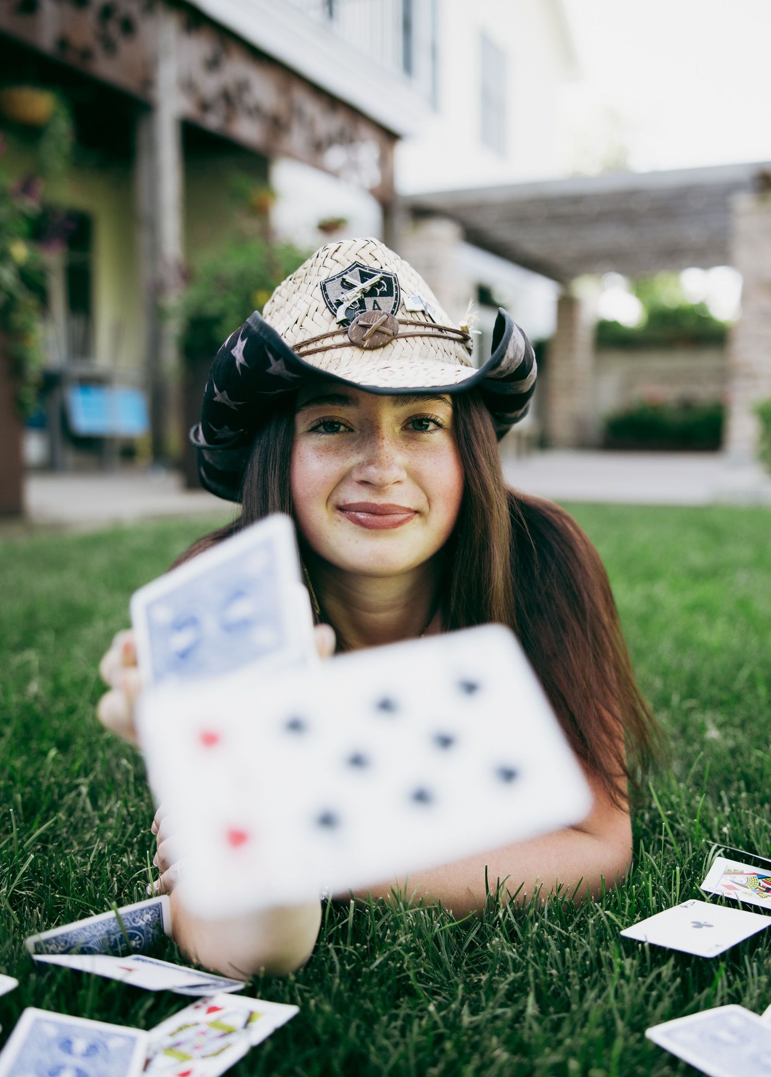 Young woman in cowboy hat lying on grass holding playing cards toward camera outdoors.