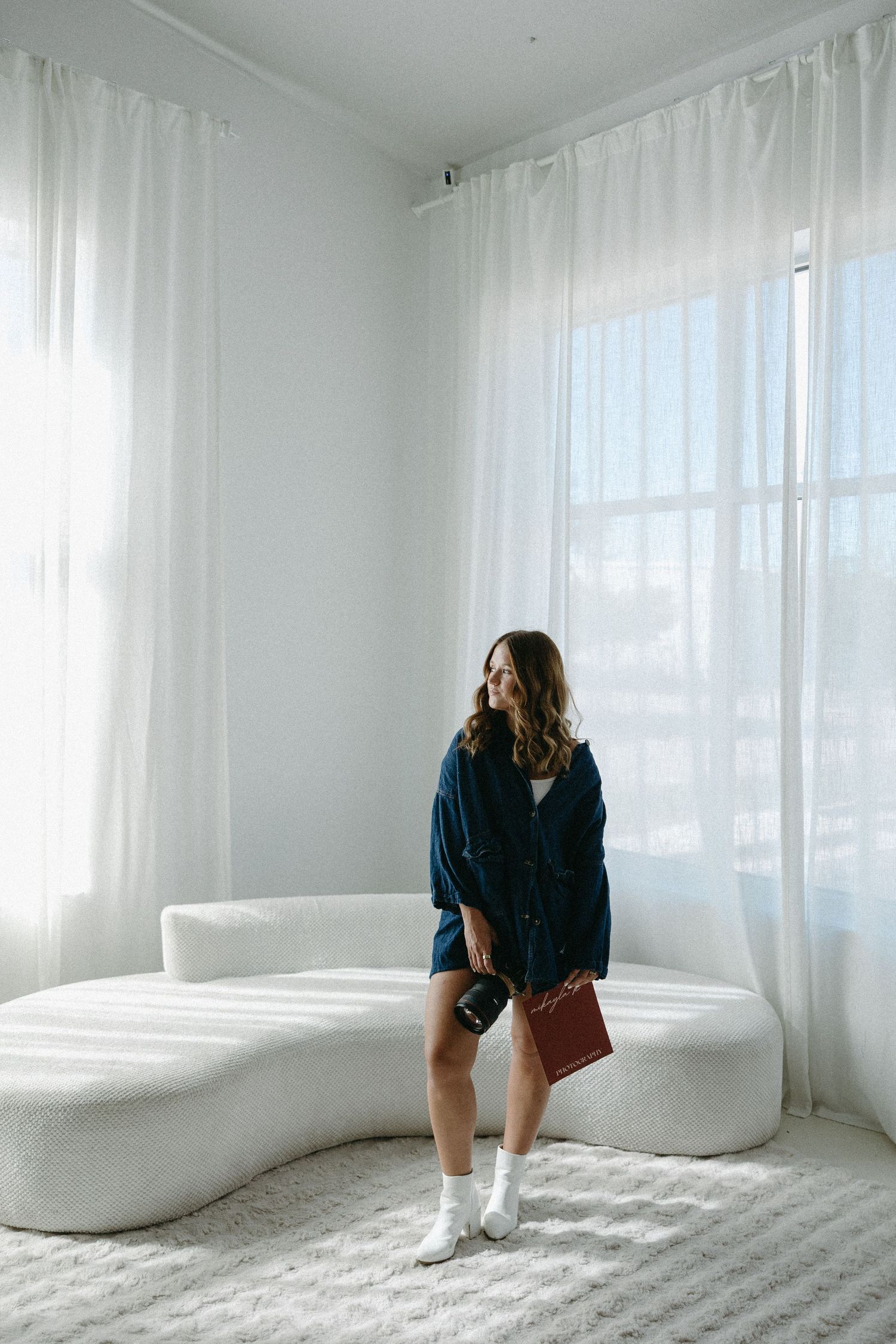 Woman in navy jacket holding brown clutch stands on white curved sofa near sheer curtains in bright room.