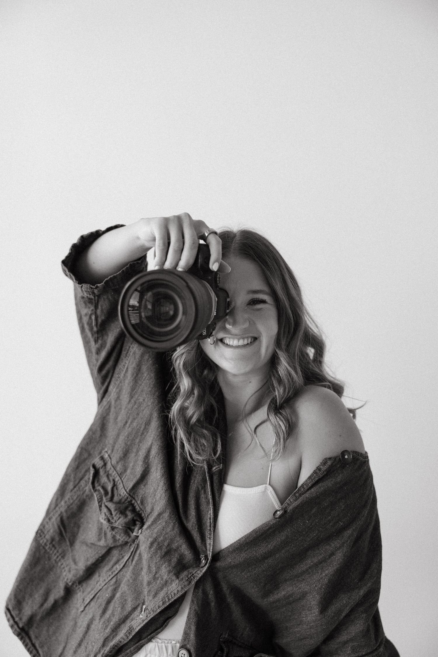 Smiling young woman holding a DSLR camera up, wearing an open jacket, in a black and white studio portrait.
