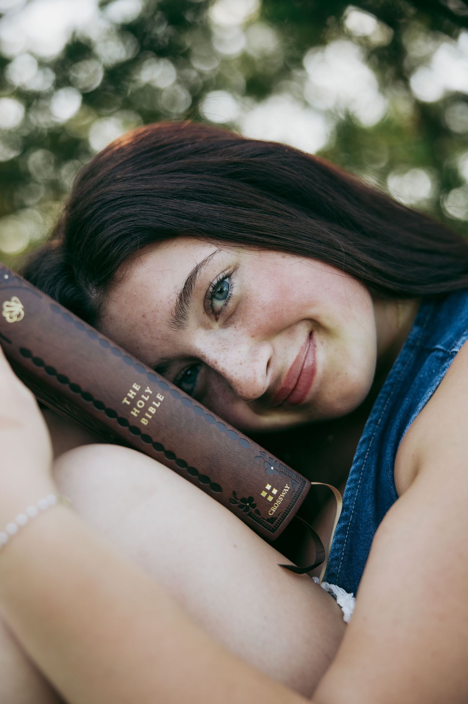 Young woman with blue eyes smiling while resting her head on a Holy Bible outdoors.