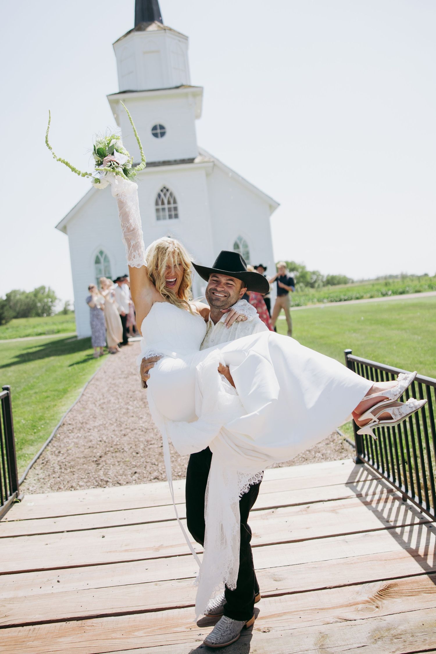 Groom carrying bride in white dress outside a white chapel on their wedding day, bride holding bouquet.