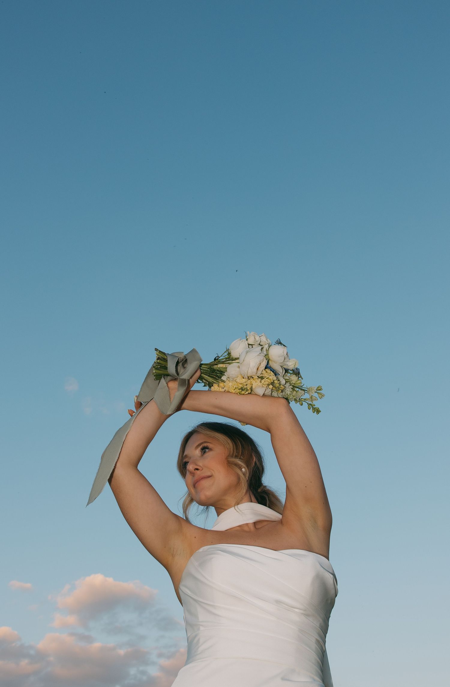 A person in a white dress holds up a bouquet against a bright blue sky. Sioux Falls, South Dakota.