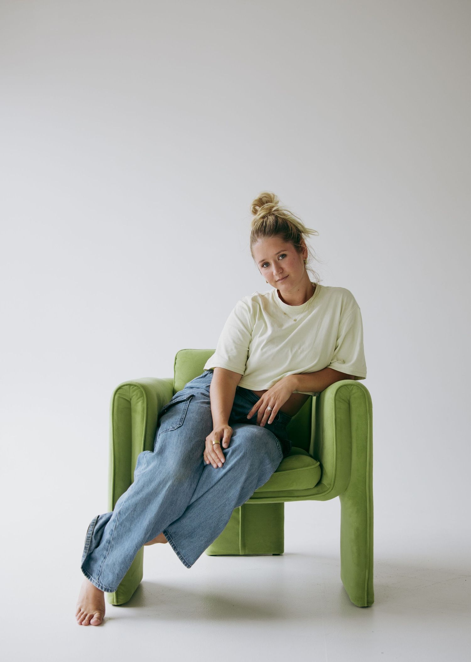 Woman in white tee and wide-leg jeans sitting relaxed on a green velvet armchair against a white background.