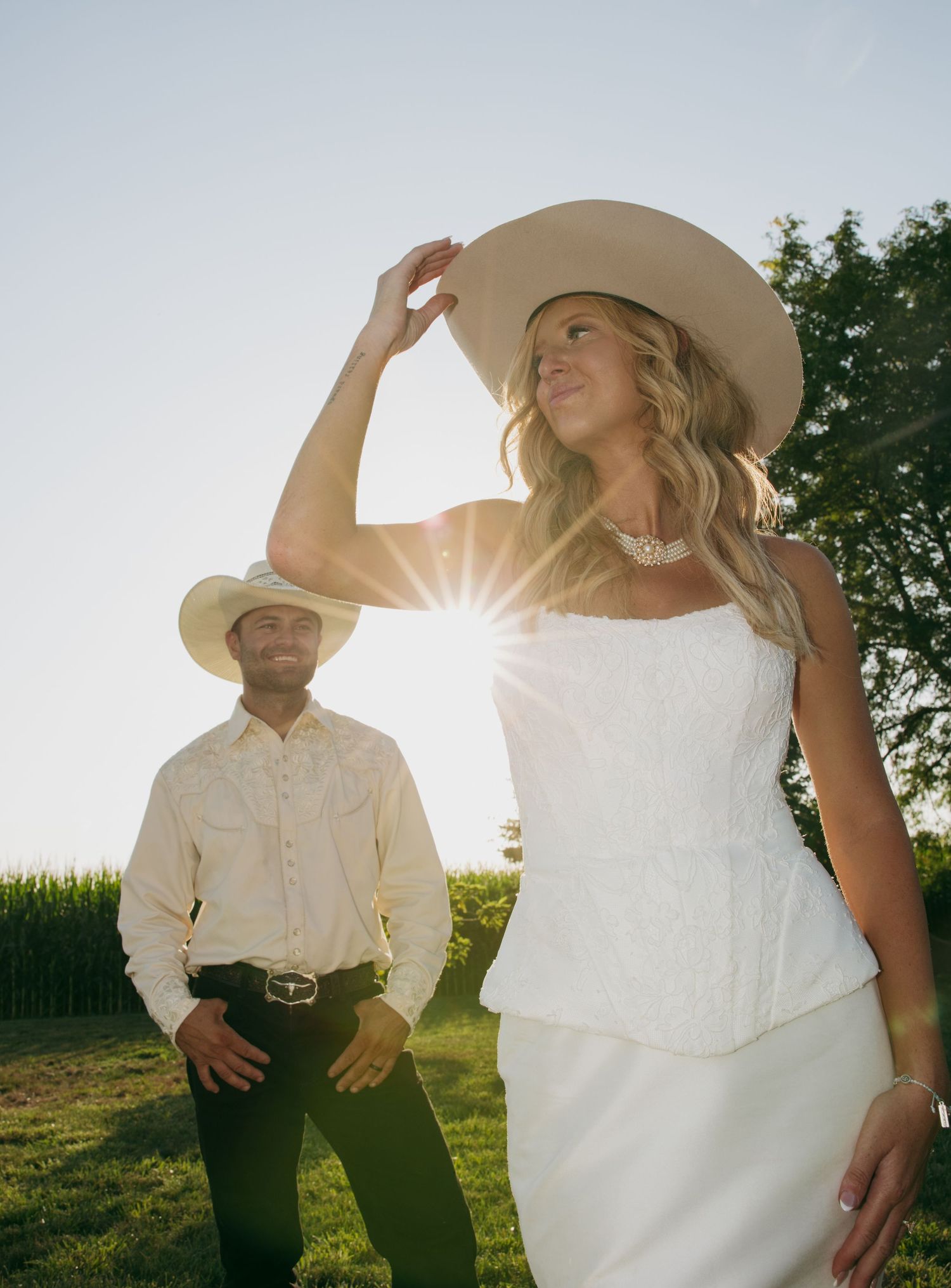 Couple in white cowboy hats poses outdoors at golden hour, bride in white peplum dress holding hat brim with sunburst.