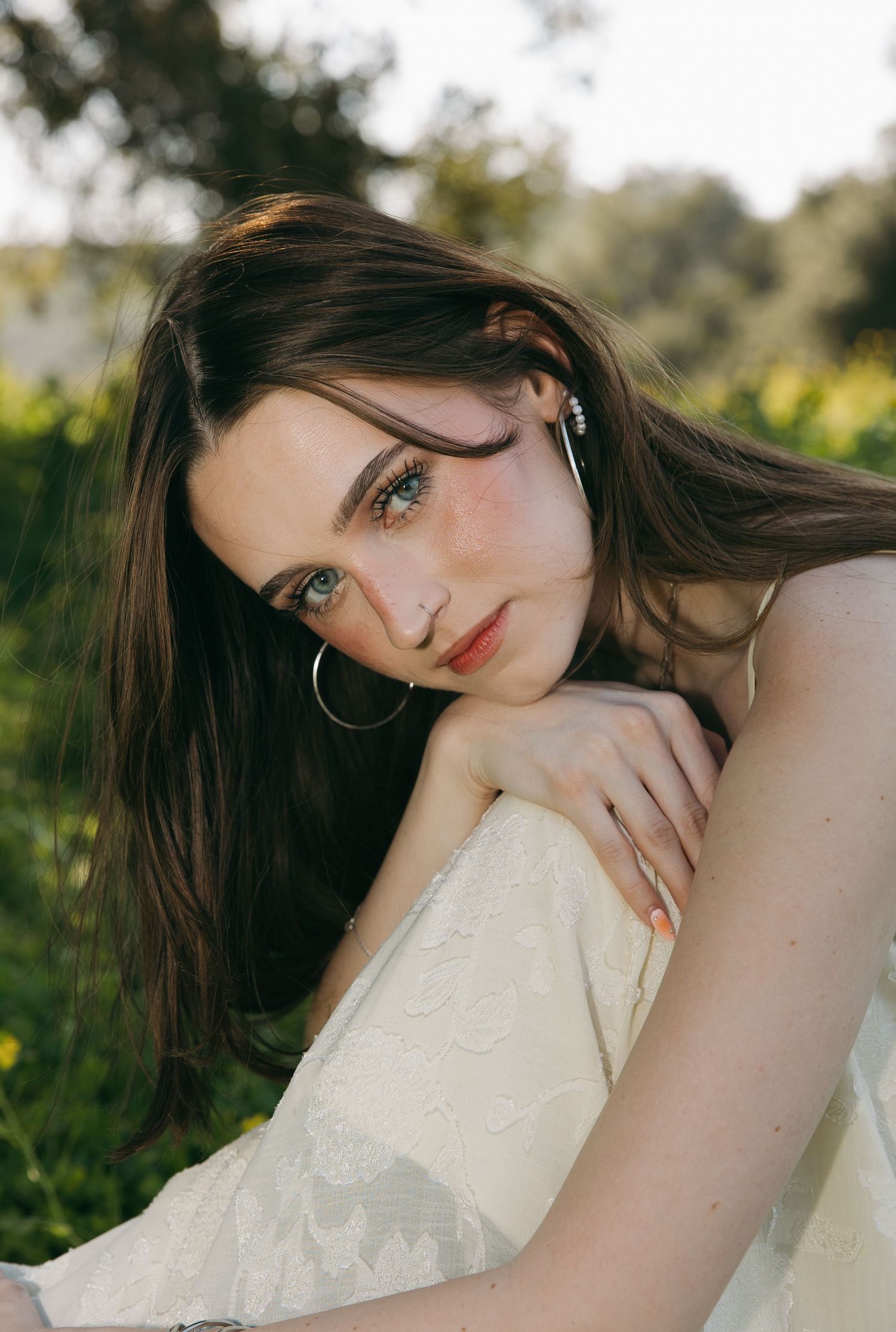 Young woman with dark hair and hoop earrings poses outdoors in white lace dress among green grass and trees.