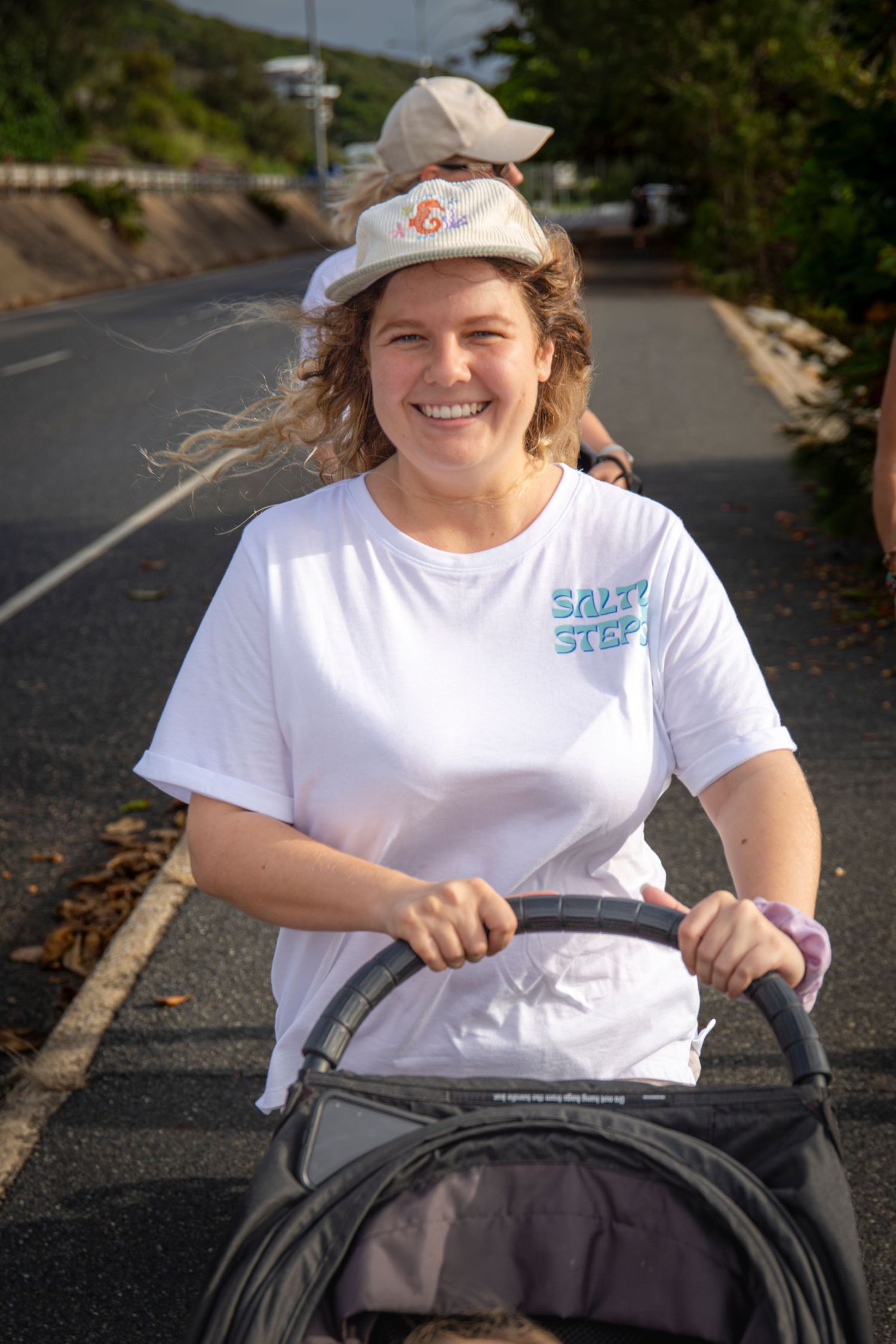 Smiling woman wearing Salty Steps t-shirt and hat pushing pram