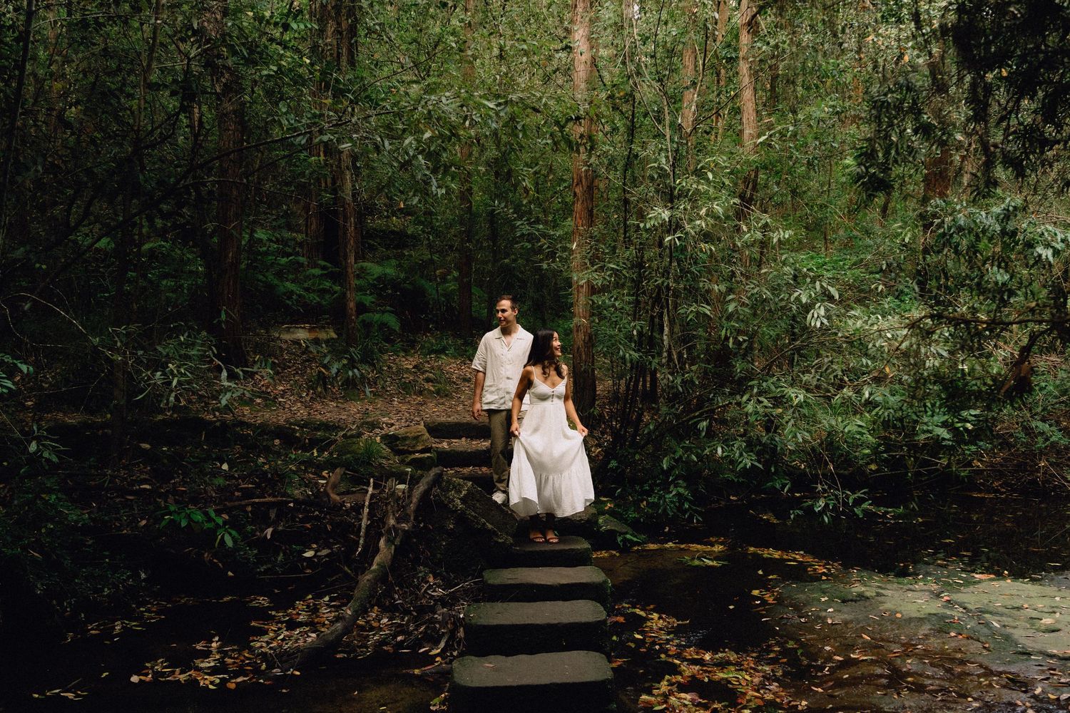 Couple in white attire walking down stone steps through a lush green forest pathway.