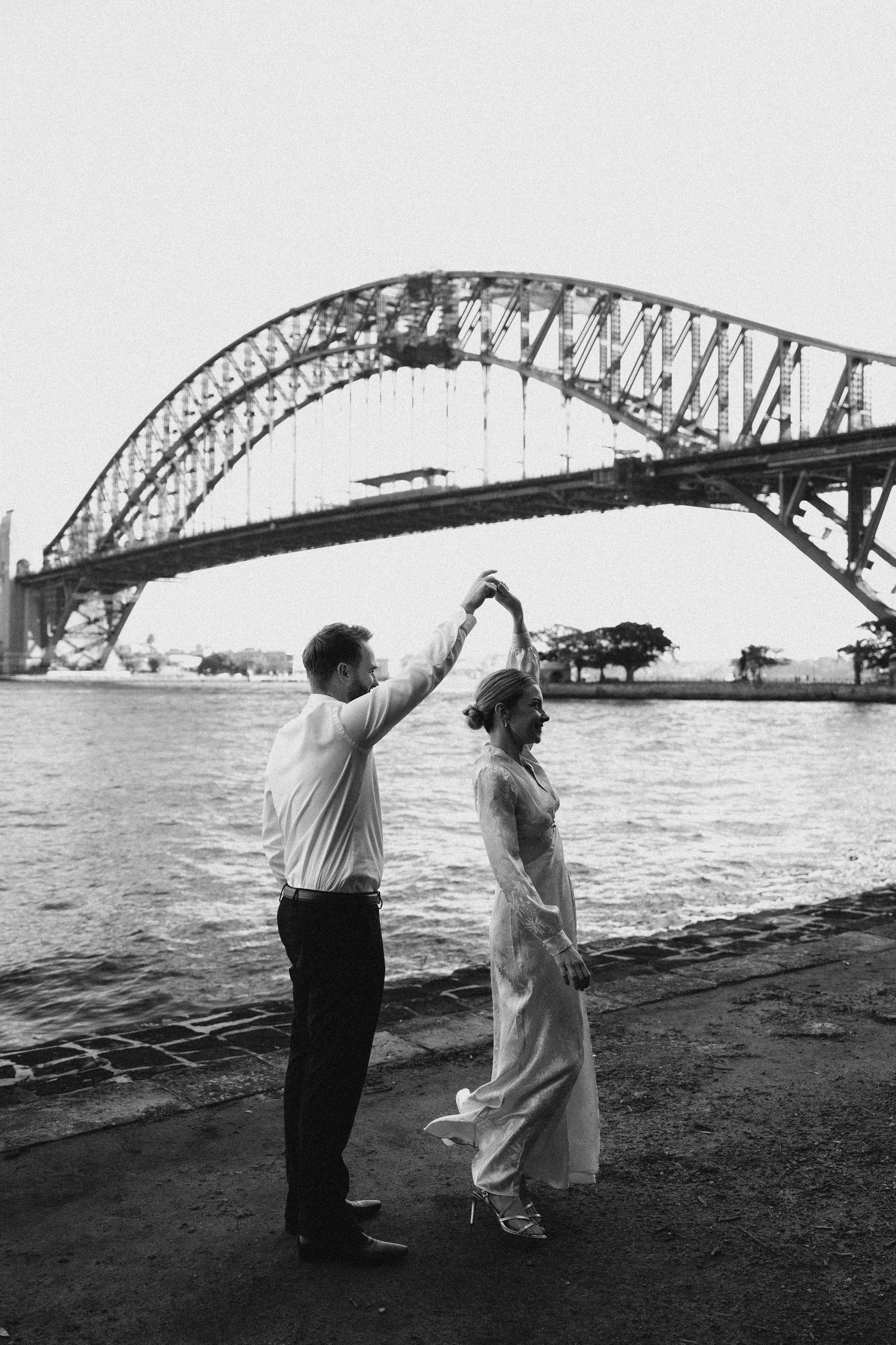 Couple dancing near Sydney Harbour Bridge waterfront in black and white wedding photo.