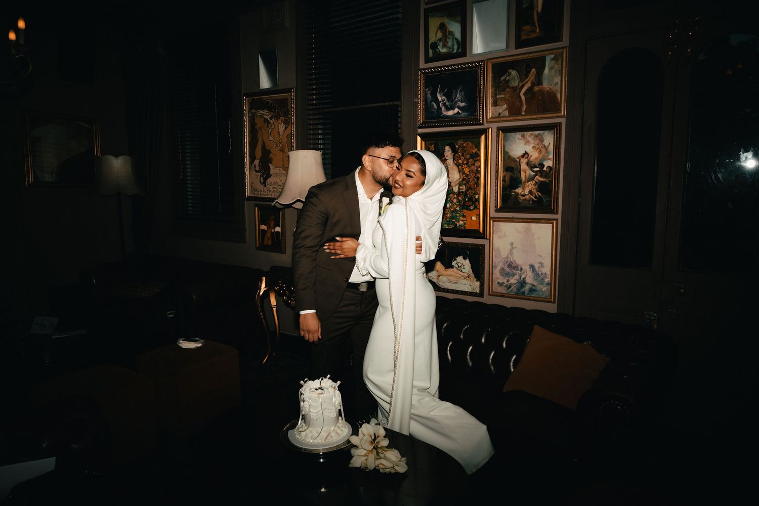 Elegant couple in wedding attire embracing in a dimly lit room with framed artwork on the wall behind them.