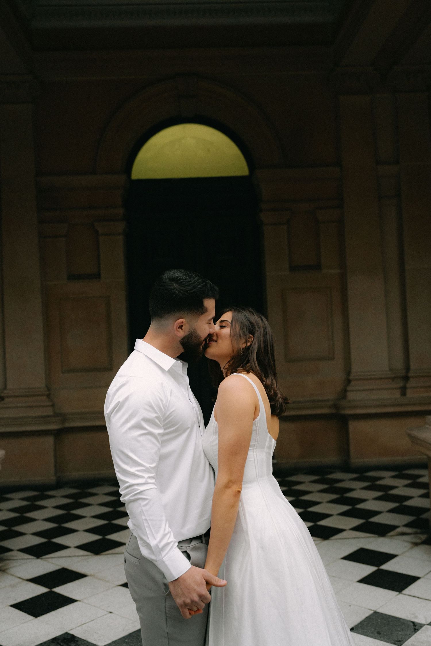 Couple sharing a romantic kiss in wedding attire on a black and white checkered floor before grand arched doorway.