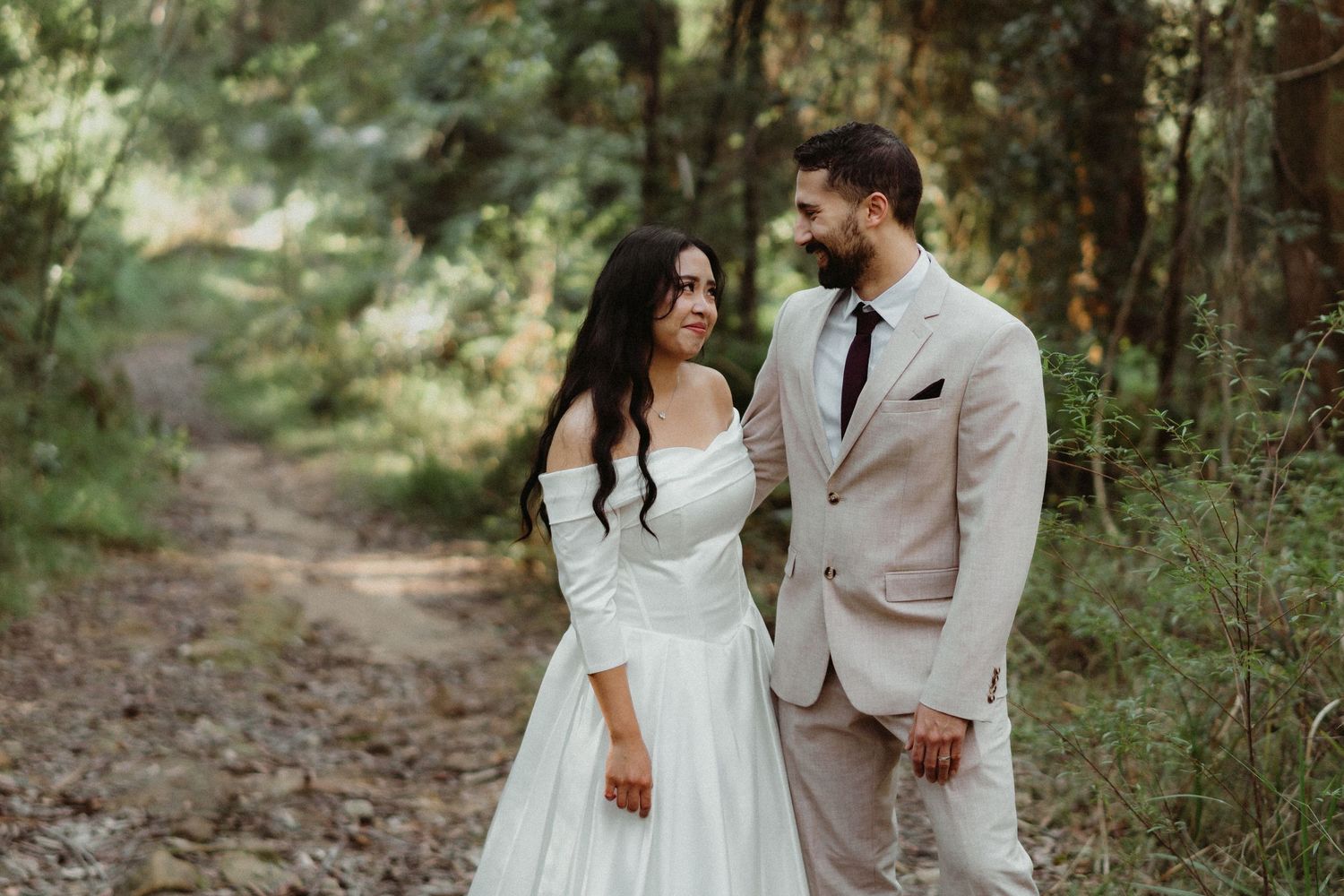 A couple in wedding attire shares intimate moments while walking through a lush green forest setting.