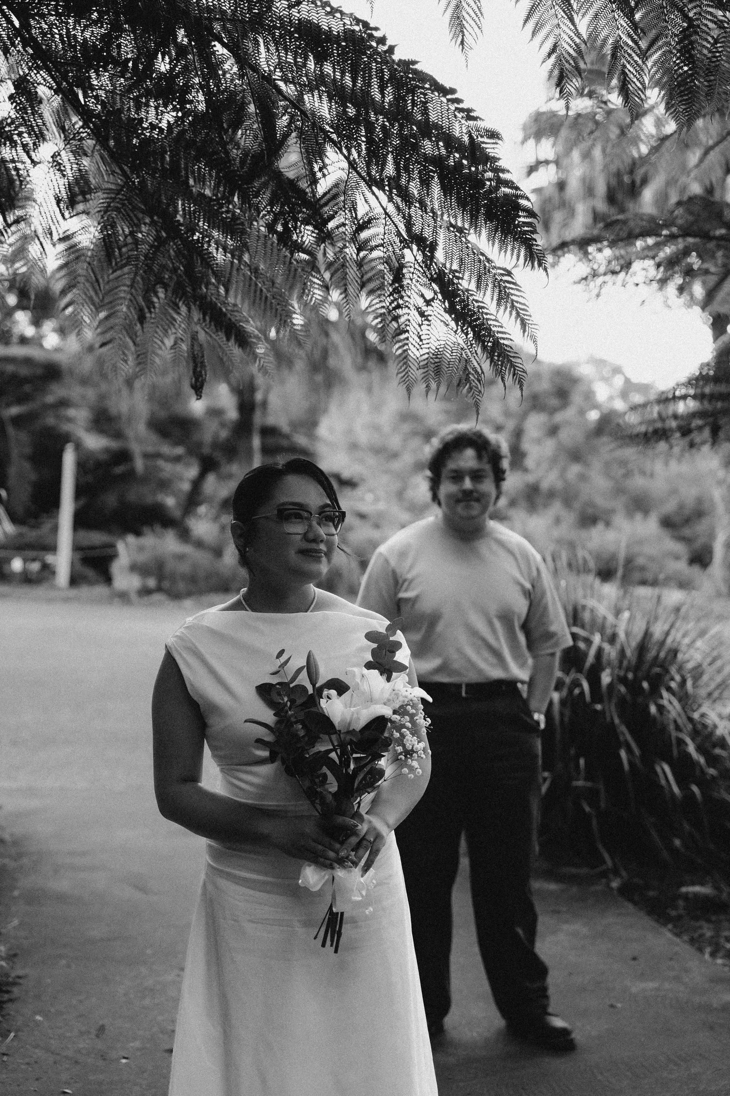 Black and white wedding photo of bride holding bouquet with groom standing behind her in a lush garden setting.