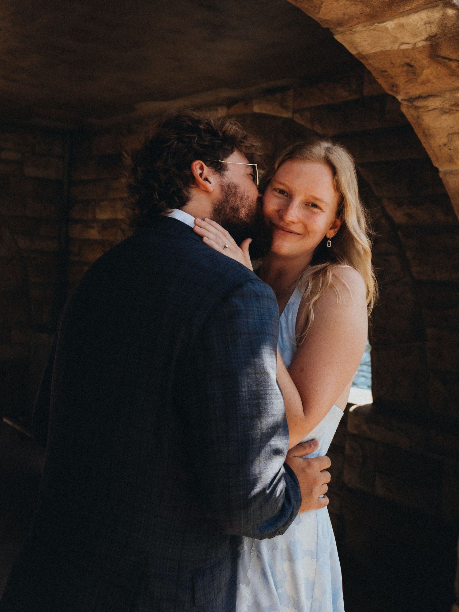 A couple embraces under a stone arch, the woman in a blue dress smiling warmly as the man nuzzles her cheek.
