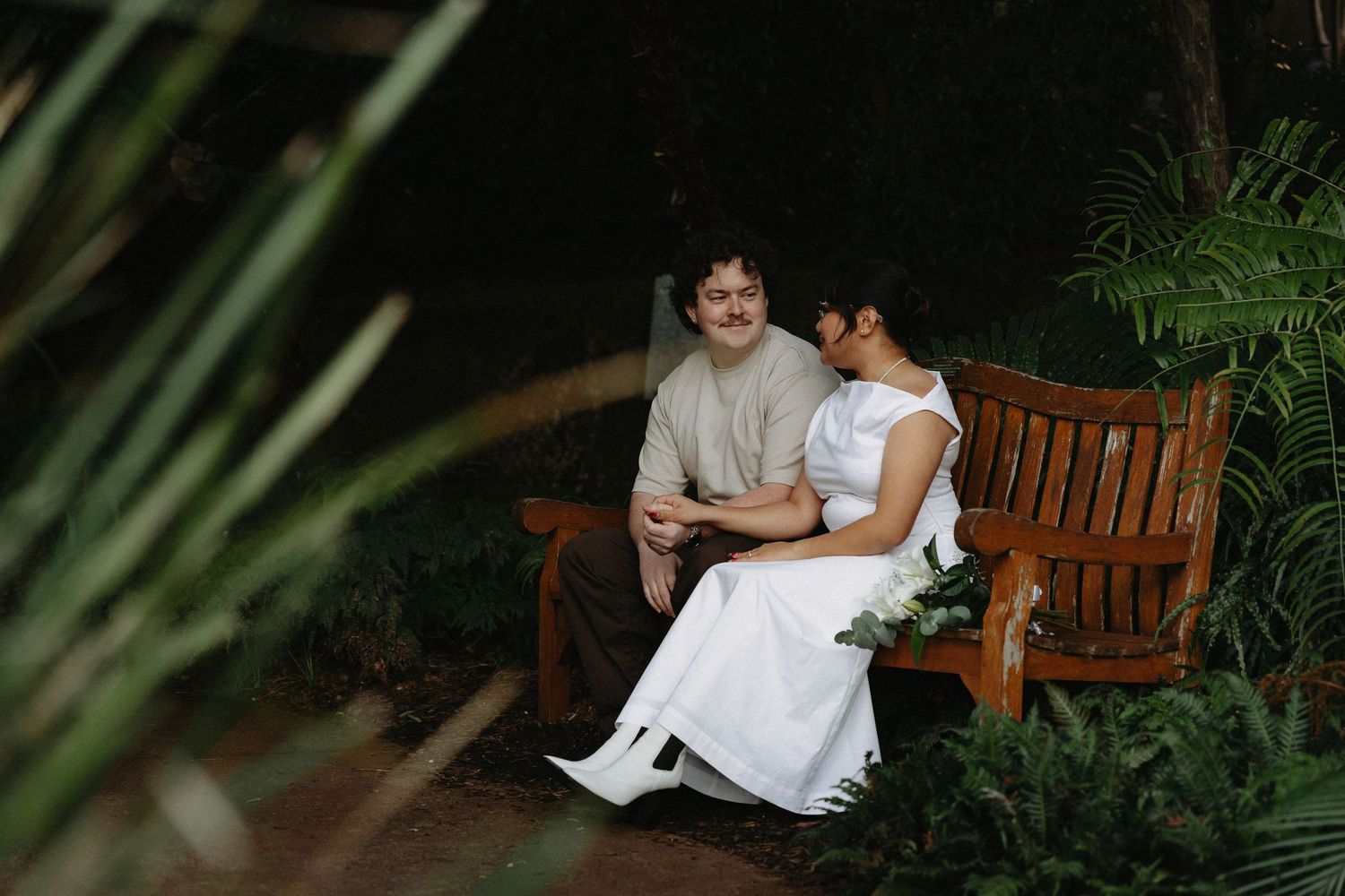 A couple in wedding attire sits together on a wooden bench surrounded by lush tropical greenery.