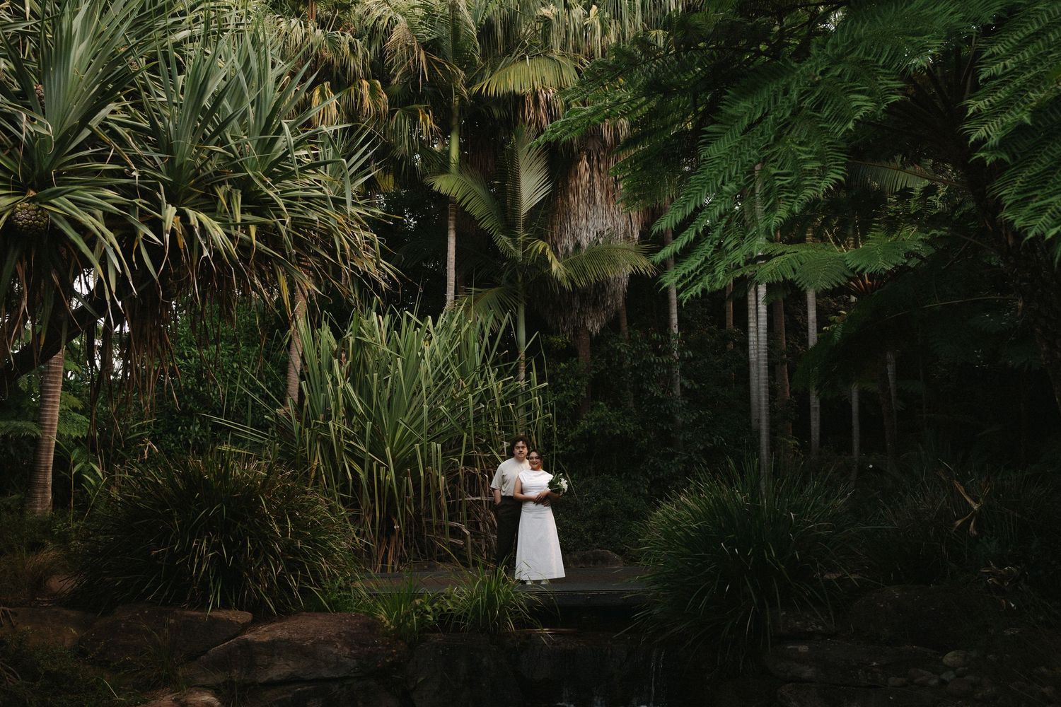 Bride in white dress holding bouquet stands on bridge surrounded by lush tropical palm trees and greenery.