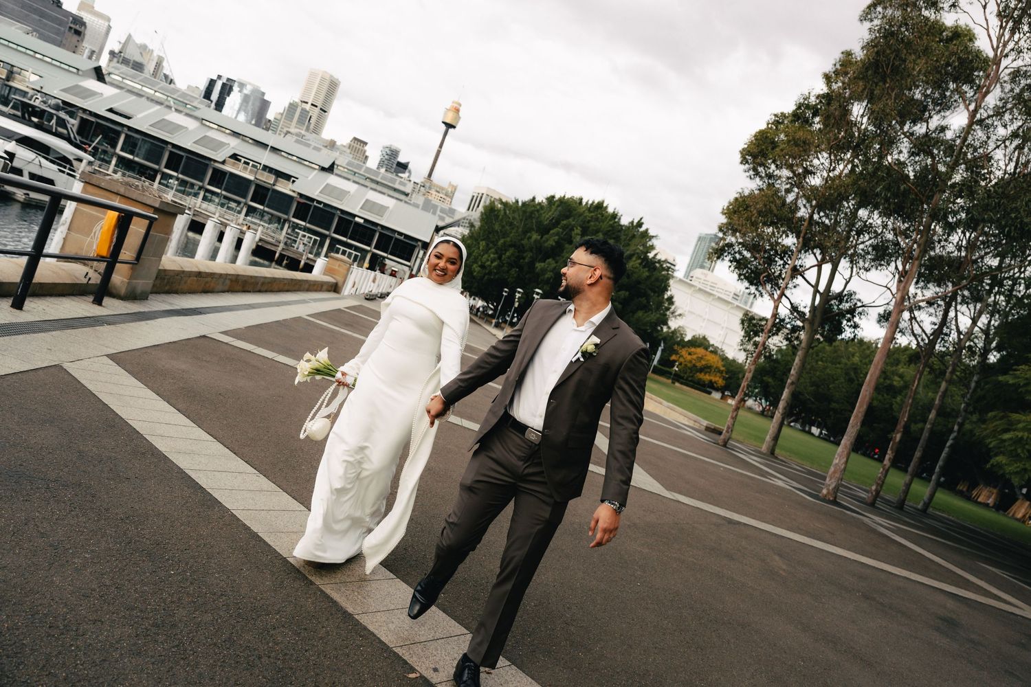 Muslim bride in white hijab and groom in grey suit hold hands walking outdoors near waterfront on their wedding day.