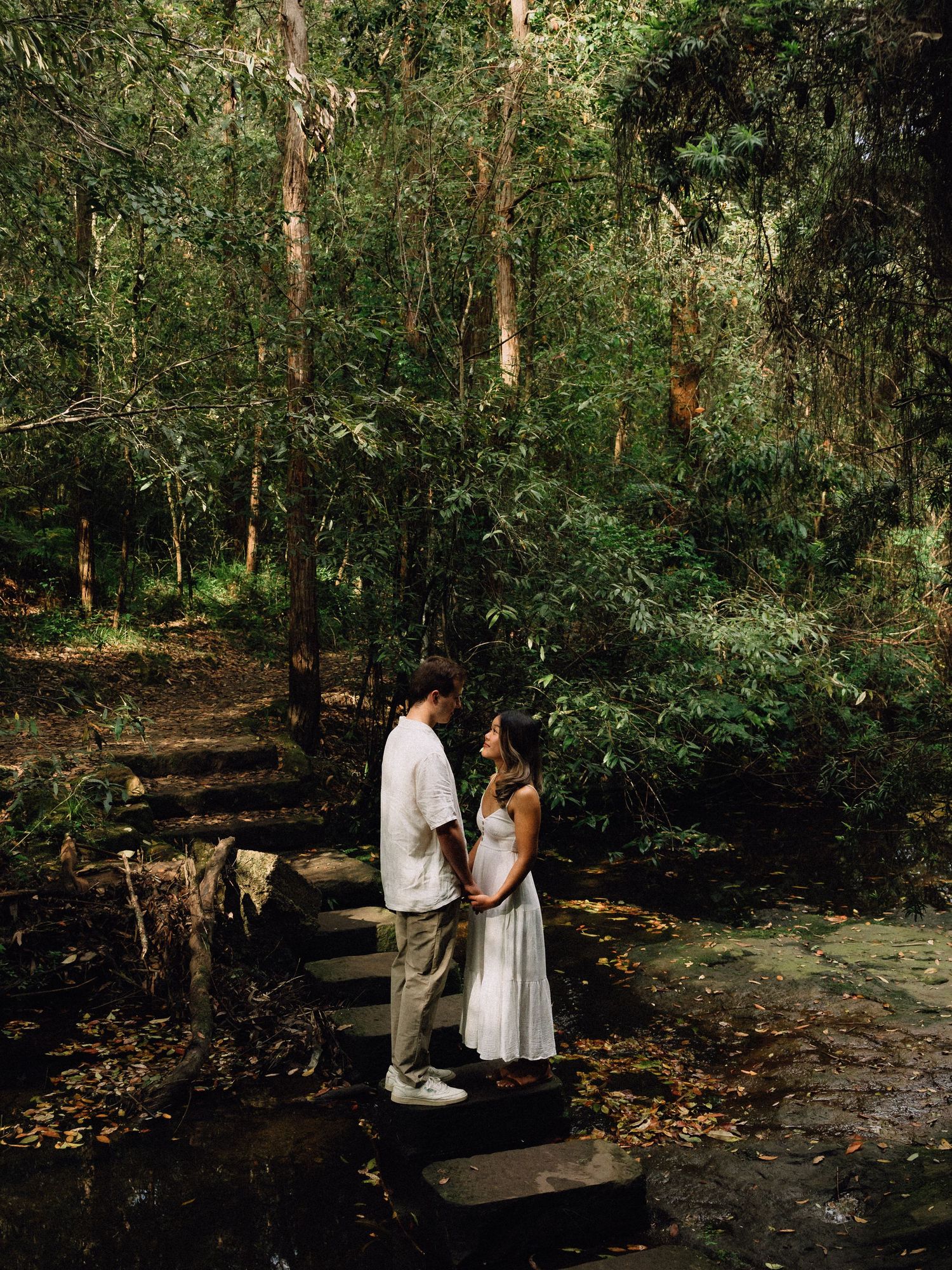 Couple holding hands on stone steps in a lush green forest, wearing white outfits in a romantic woodland setting.