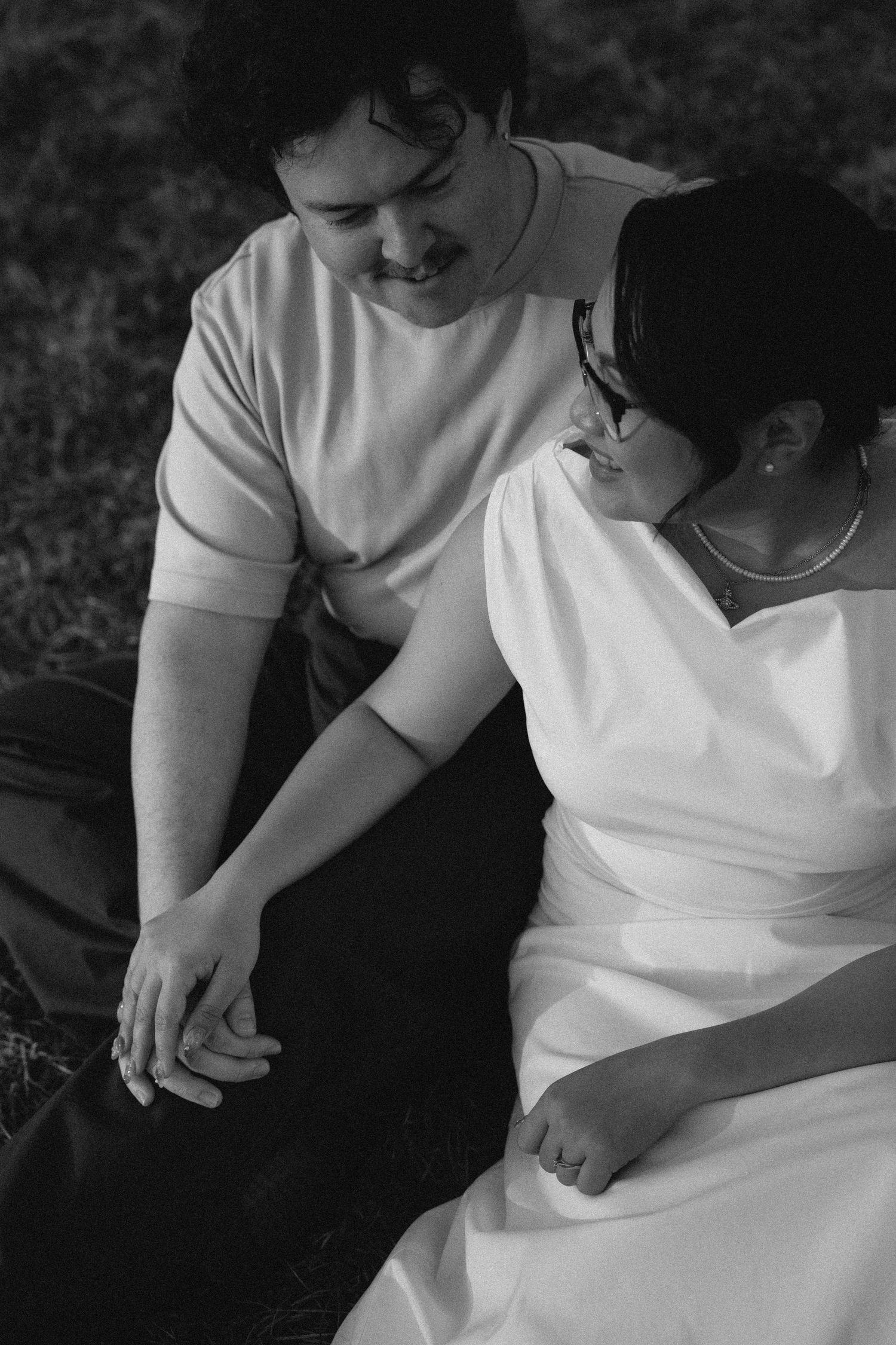 Black and white photo of a smiling couple holding hands while sitting on grass, sharing a tender moment.