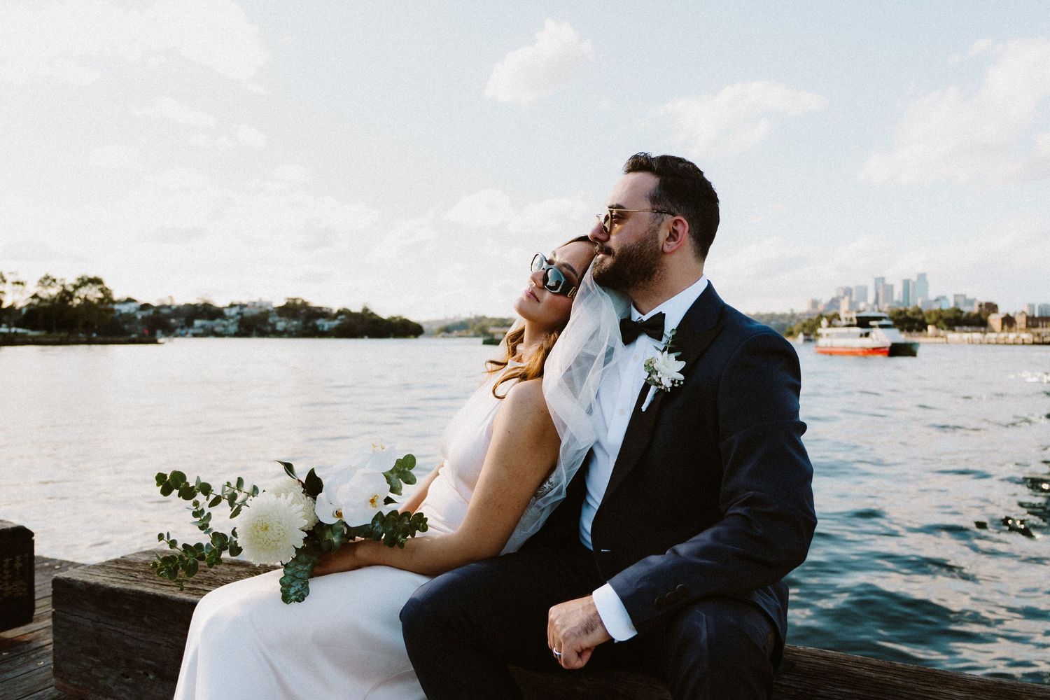 Bride and groom sitting on a waterfront dock, sharing a romantic moment with city skyline and harbor in the background.