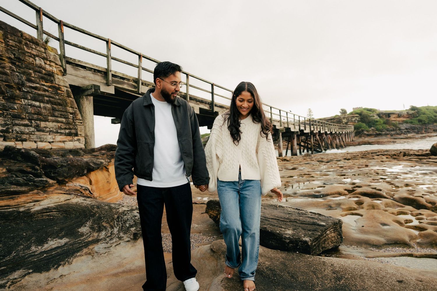 Couple holding hands walking along rocky coastal shoreline near a wooden bridge on an overcast day.