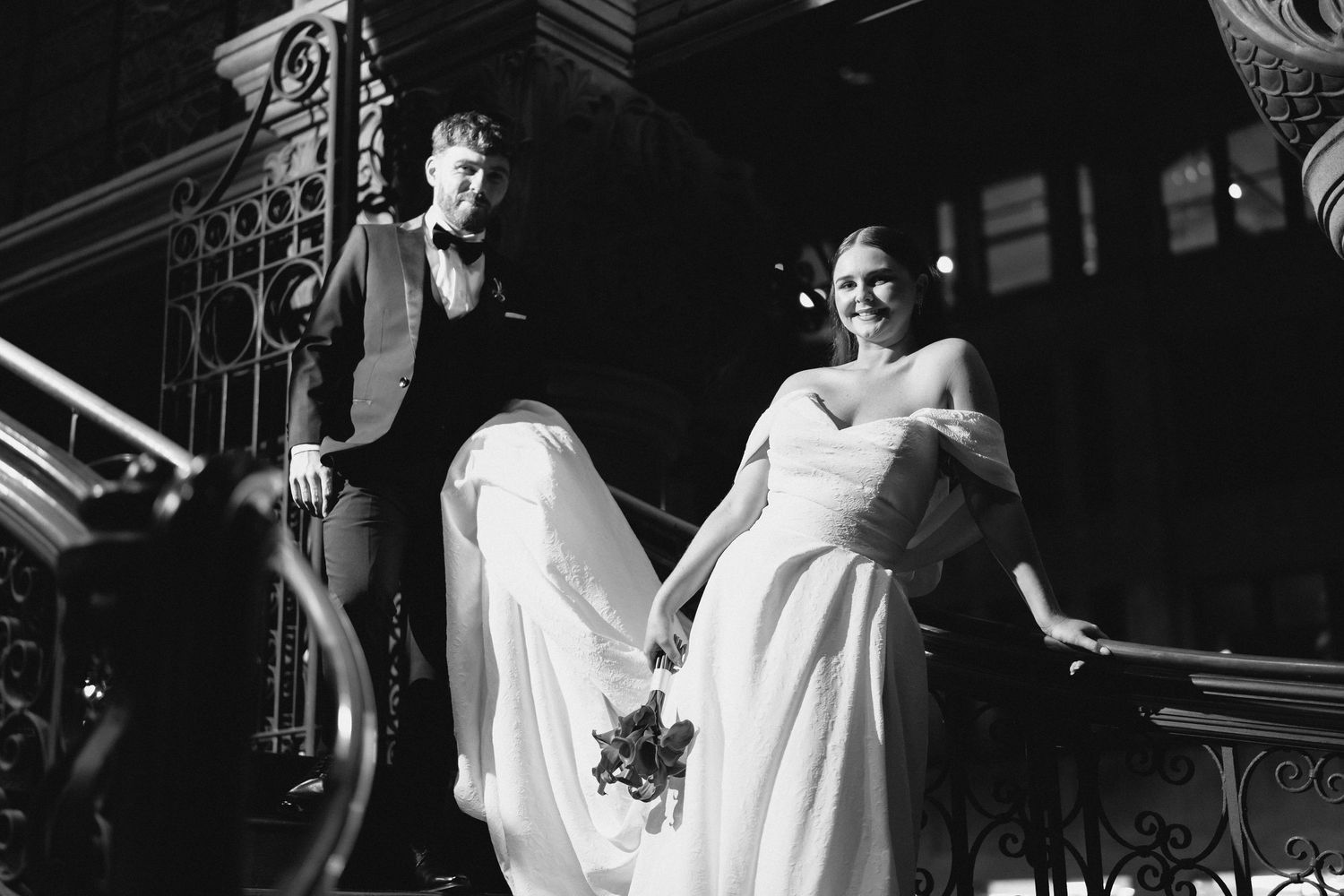 Black and white wedding photo of bride and groom on elegant staircase, bride holding dress and bouquet.