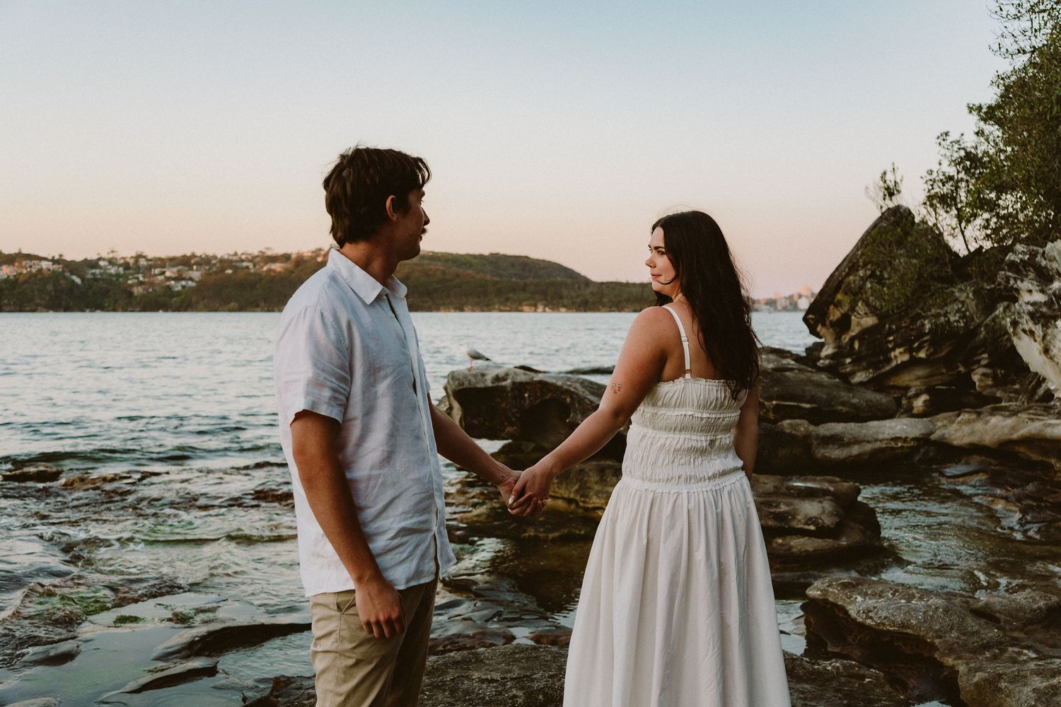 Couple holding hands on rocky shoreline at sunset, man in white shirt and woman in white dress.