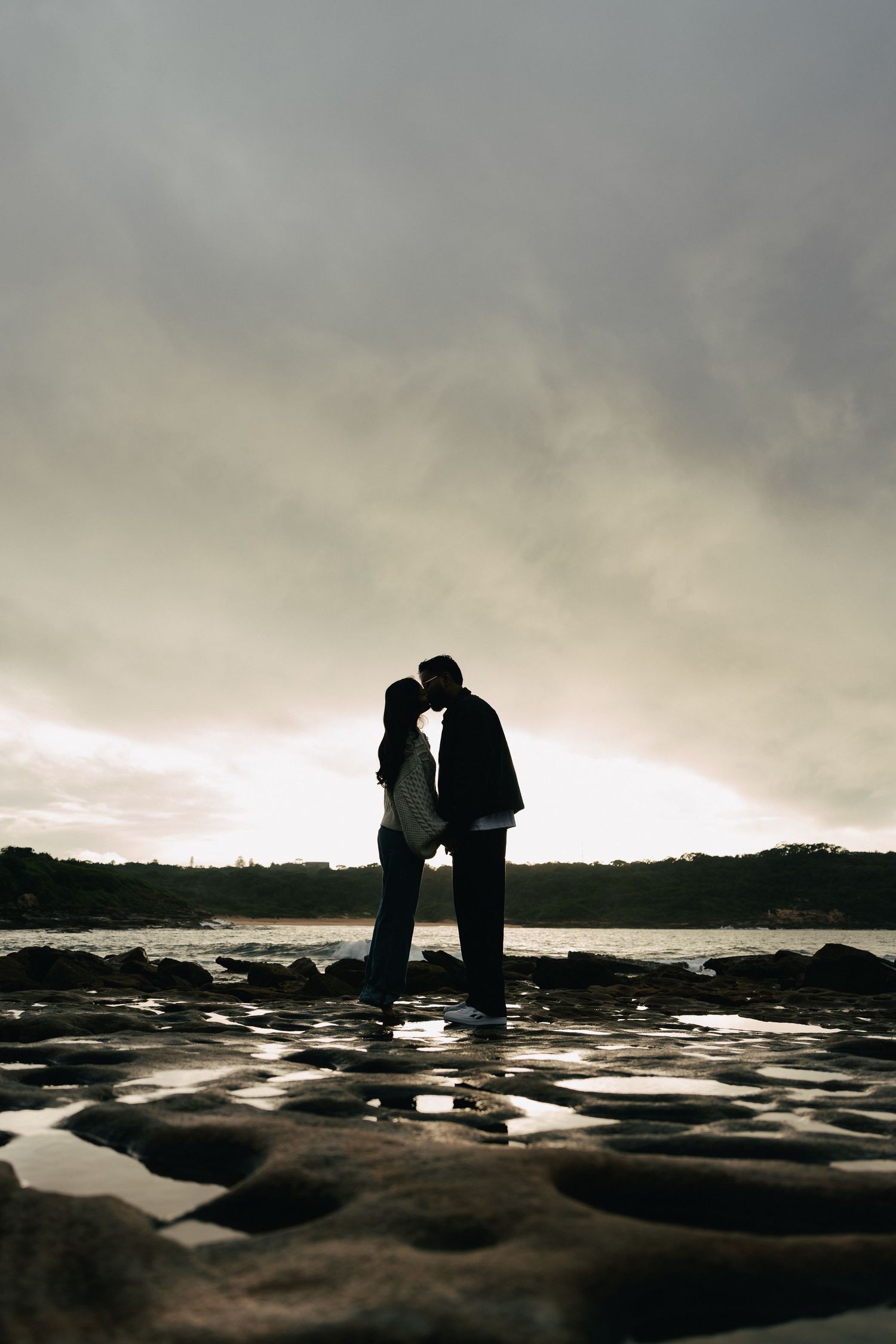 Silhouette of a couple kissing on rocky shoreline with dramatic cloudy sky and water reflections at sunset.