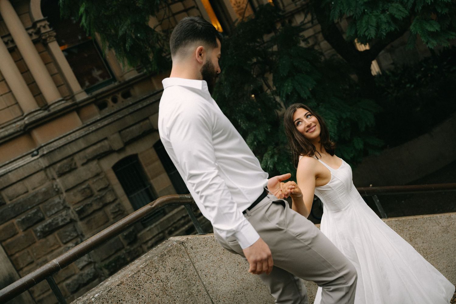 A bride and groom share a romantic moment on stone steps, dressed in white wedding attire, with lush greenery behind them.