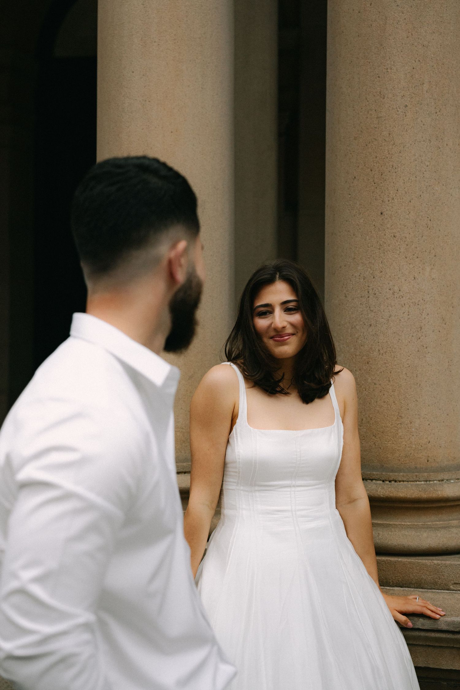 Bride in white dress smiling at groom near stone columns during wedding photo session.