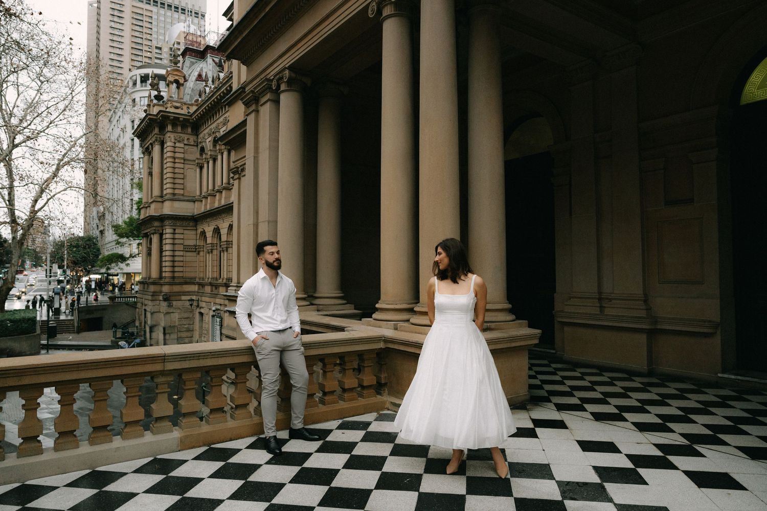 Couple in wedding attire posing on checkered floor of historic stone building with grand columns.