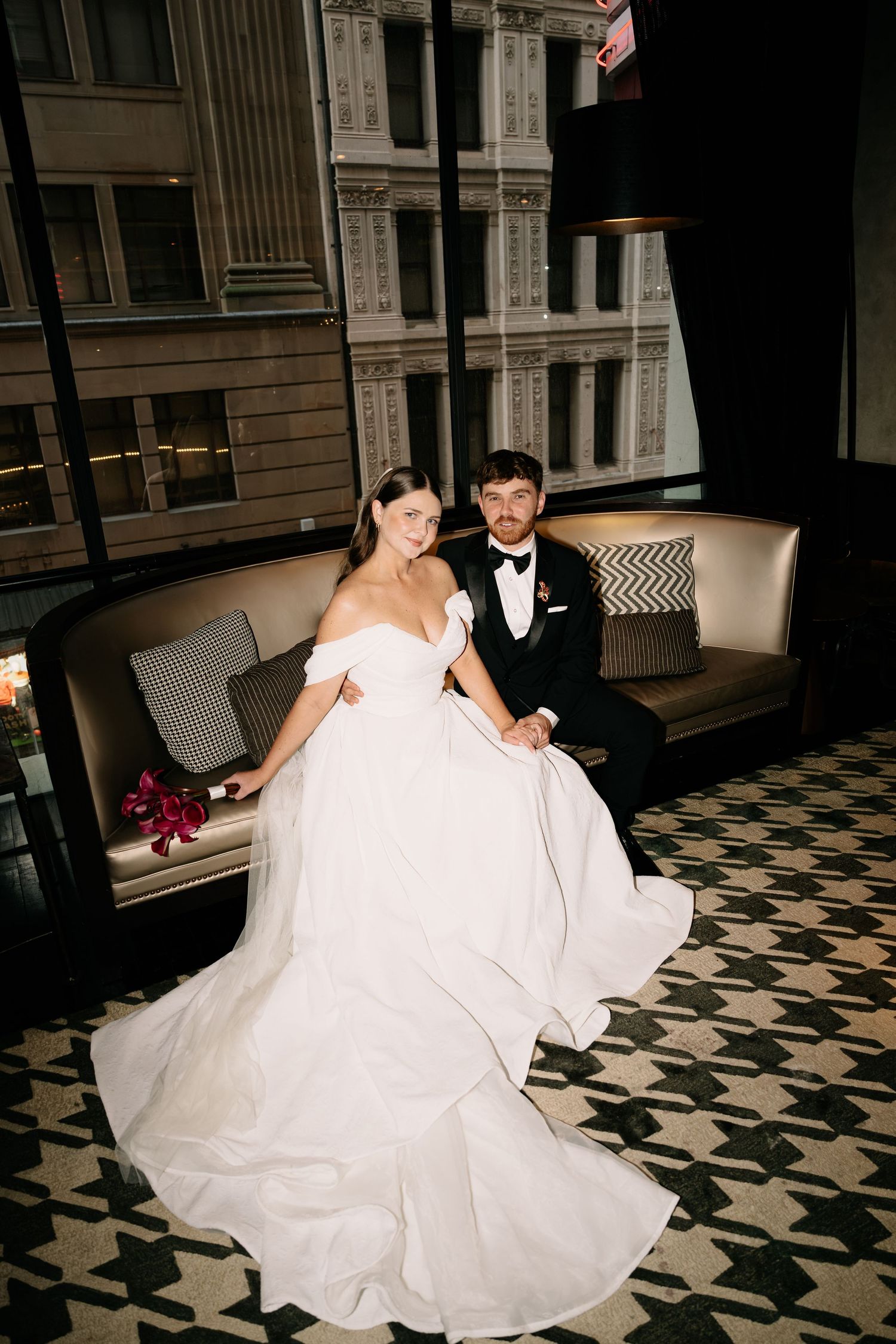 Bride in off-shoulder white gown and groom in black tuxedo sit together on a sofa in an elegant venue.