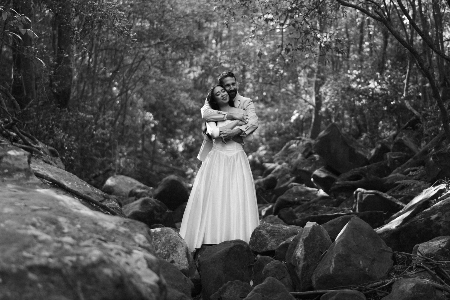 A romantic black and white photo shows a couple embracing on rocky terrain surrounded by lush forest scenery.
