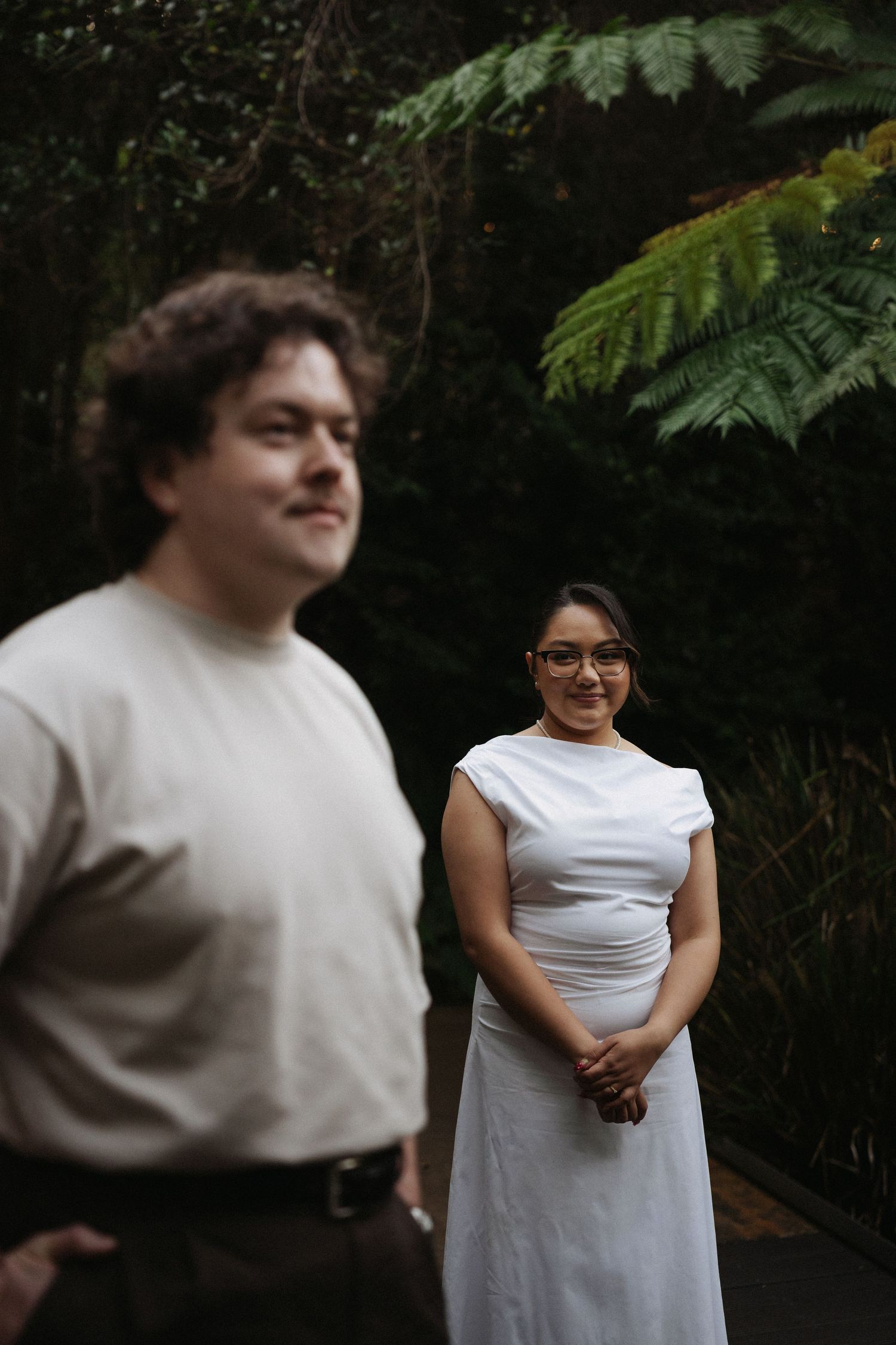 A woman in a white dress smiles in a lush green garden while a man in a beige shirt stands in the foreground.