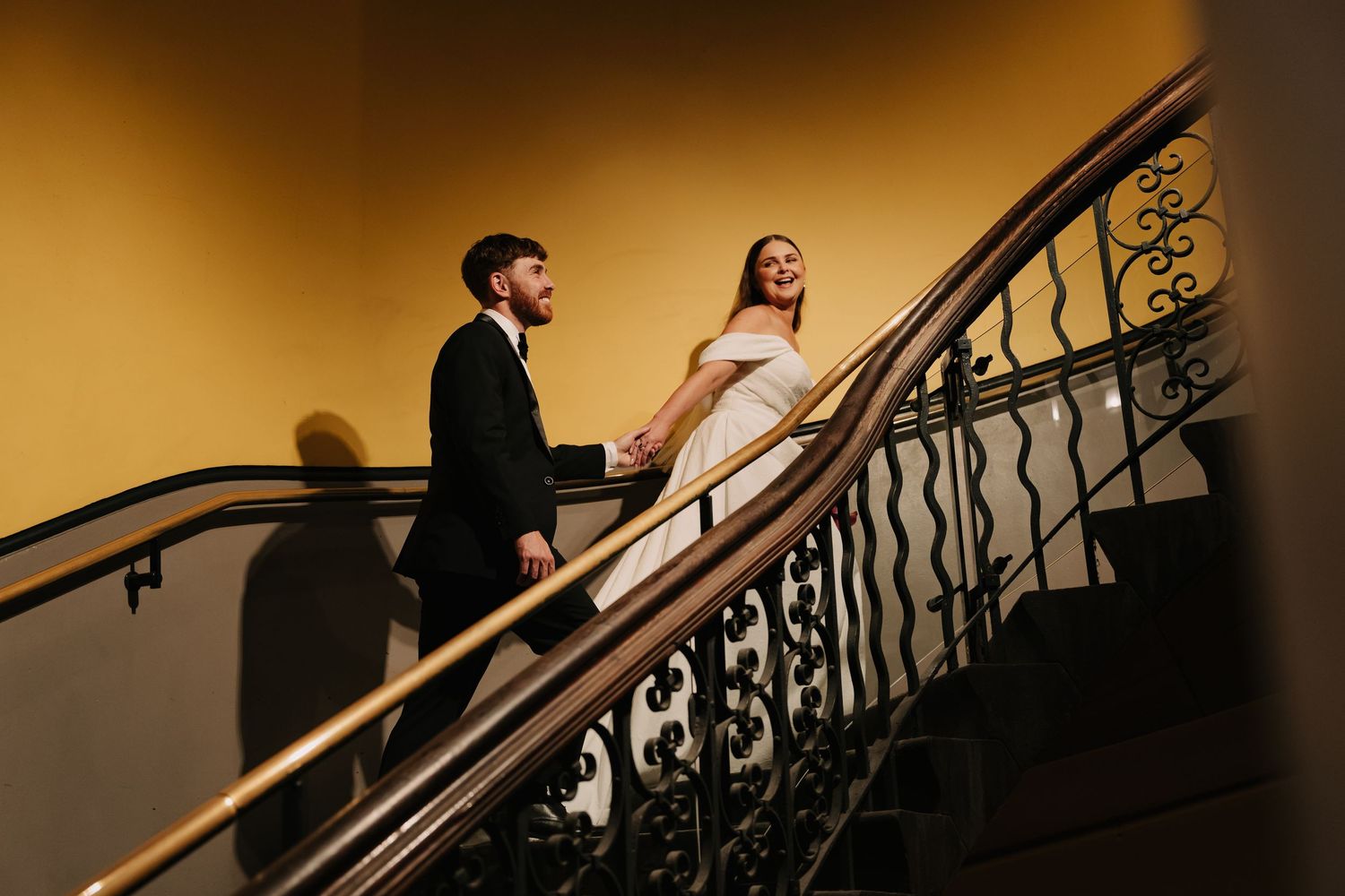 Bride and groom walk up elegant staircase with ornate railing, bathed in warm golden light.