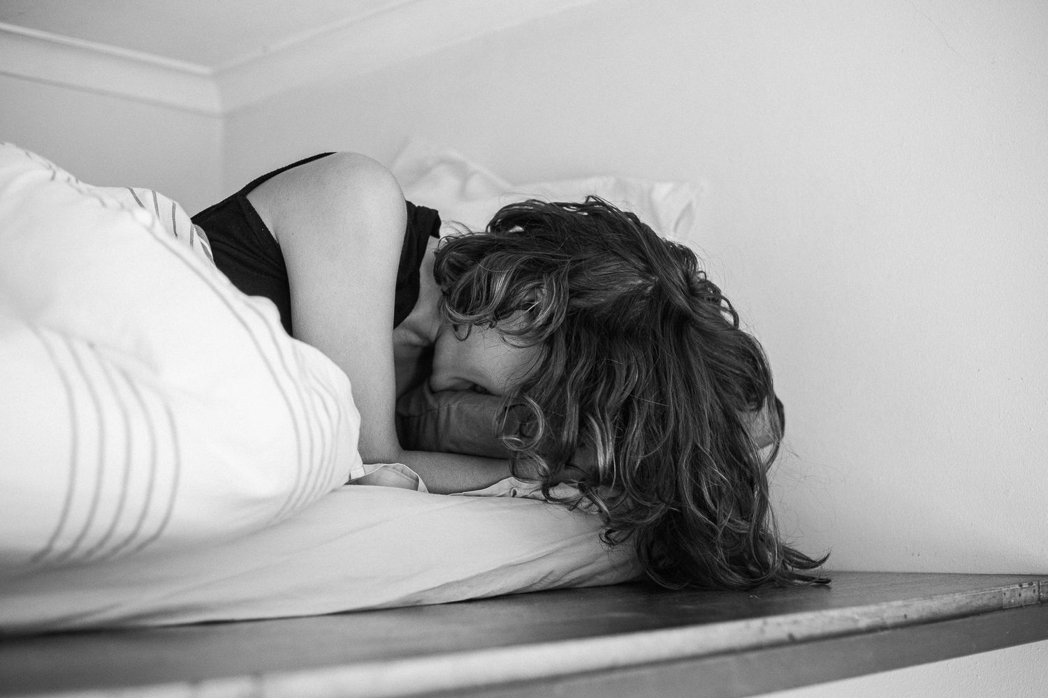 Black and white photograph showing a figure lying down in bed with white sheets and pillows.