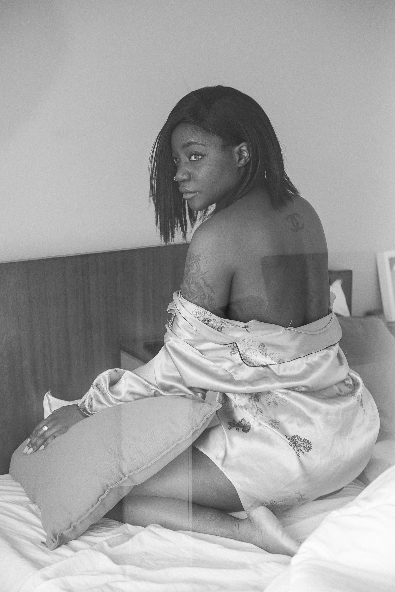 Black and white artistic portrait of someone in a silk robe sitting on a bed looking over their shoulder.