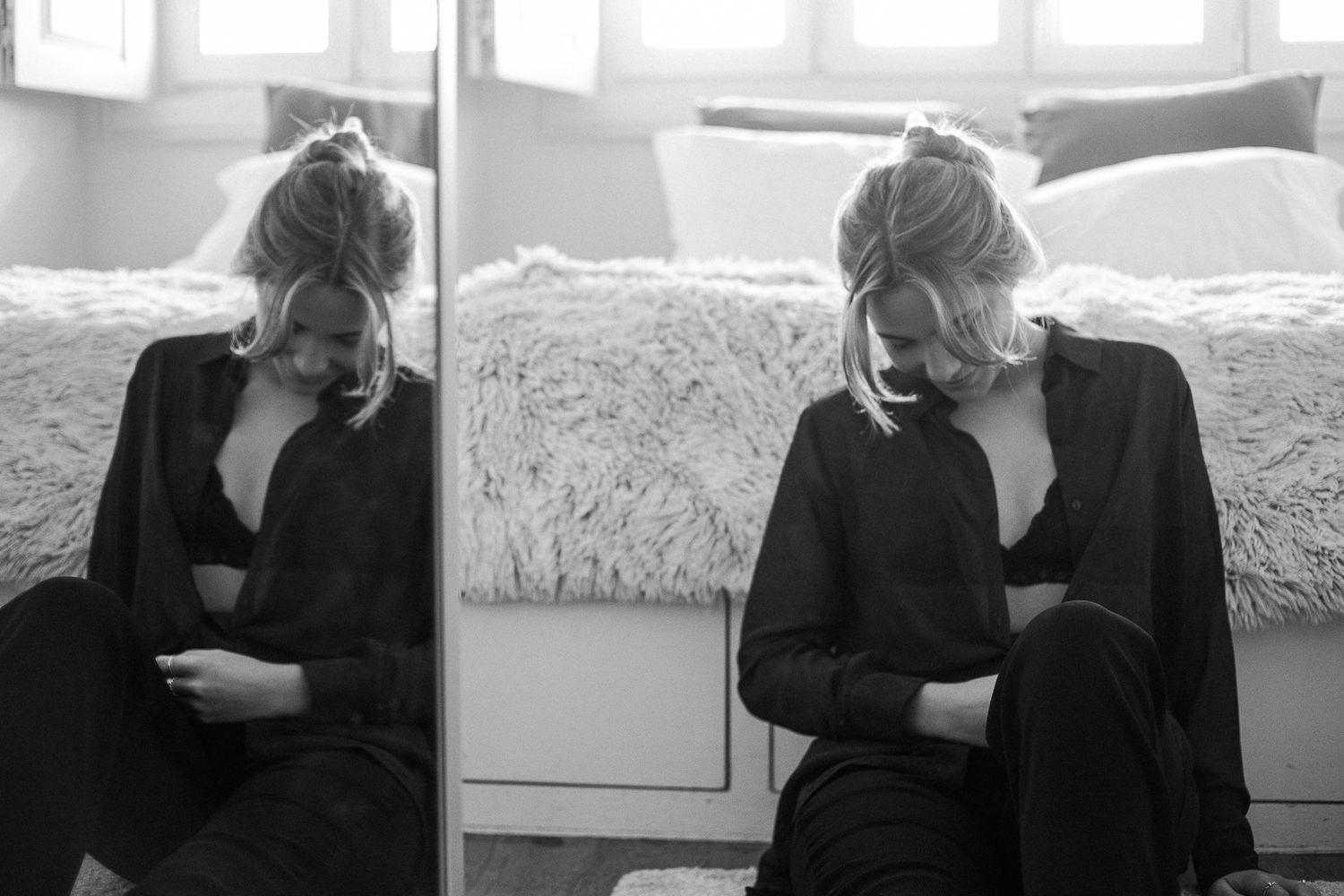 Black and white artistic portrait of person in dark clothing seated by window with moody lighting and dramatic composition.