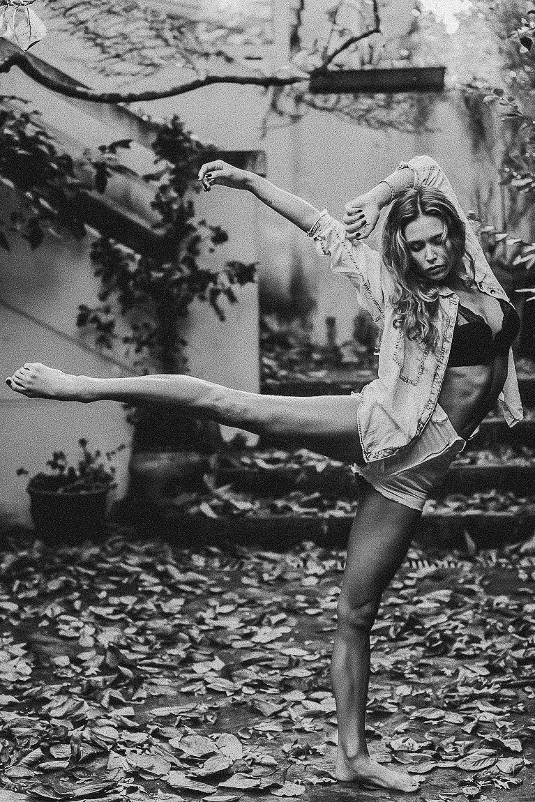 A dancer performs a graceful high kick pose amidst fallen autumn leaves in a black and white photograph.