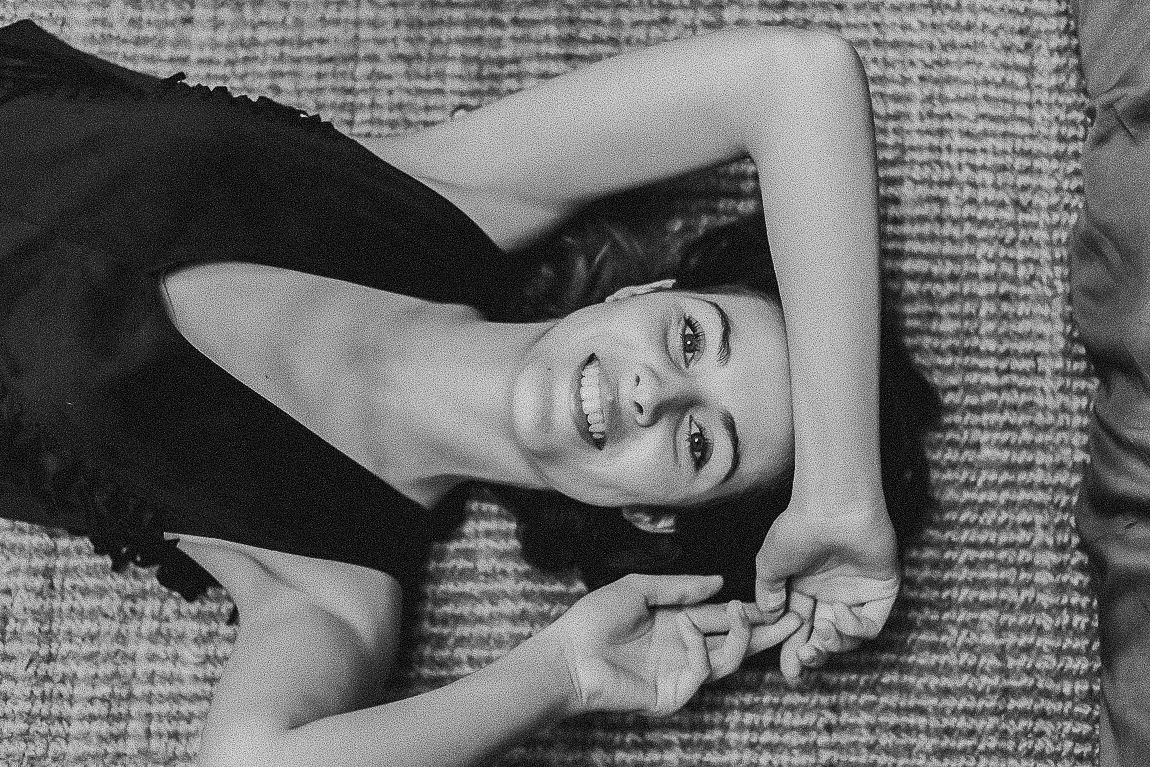 Black and white portrait of someone lying on newspaper print, smiling and looking up while wearing a black top.