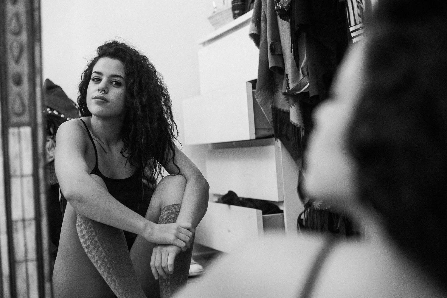 Black and white photo of someone with curly hair leaning against a window frame in dramatic lighting.